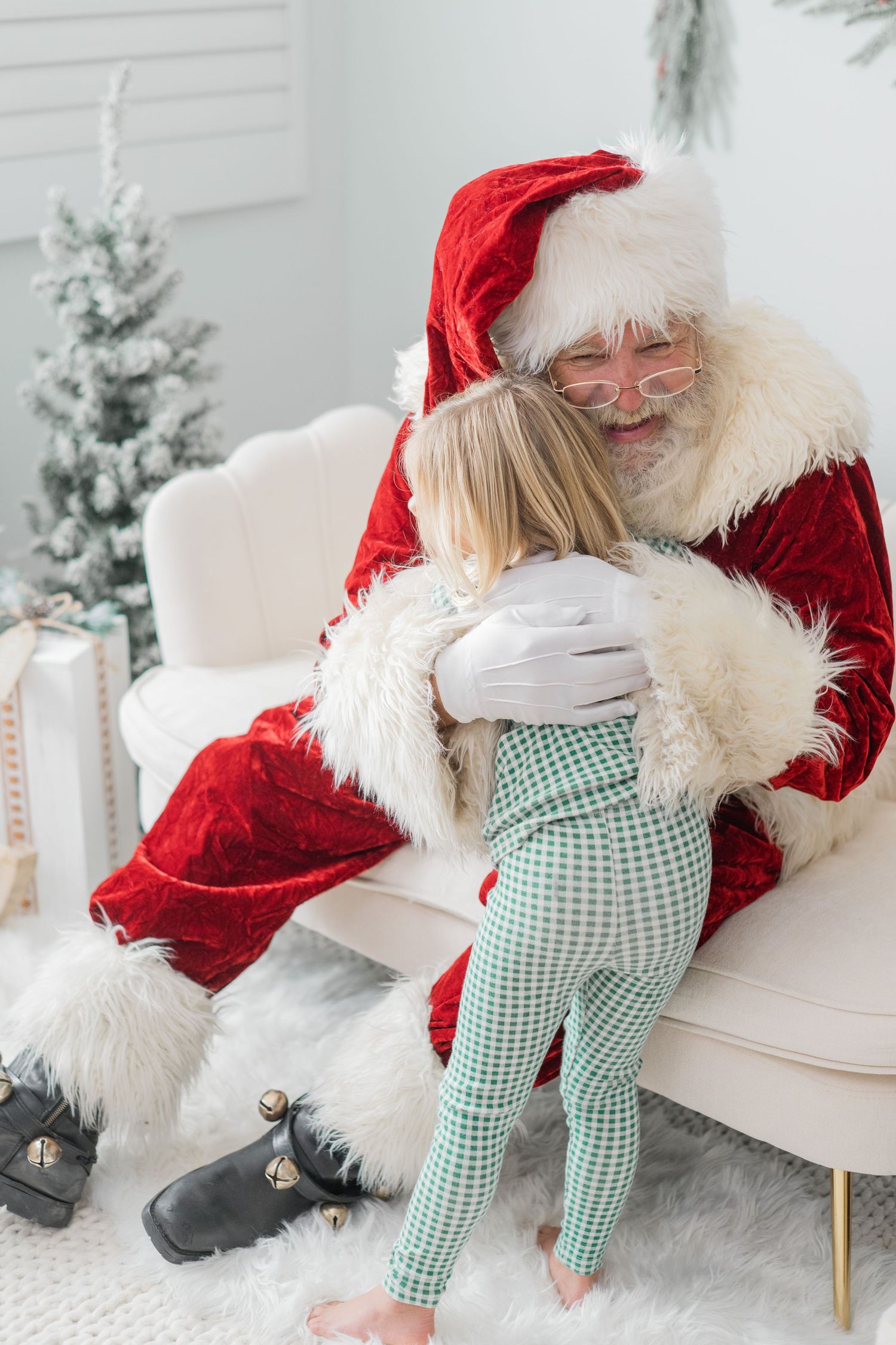 A young child in green plaid pajamas hugs Santa Claus sitting in a white chair near a Christmas tree.