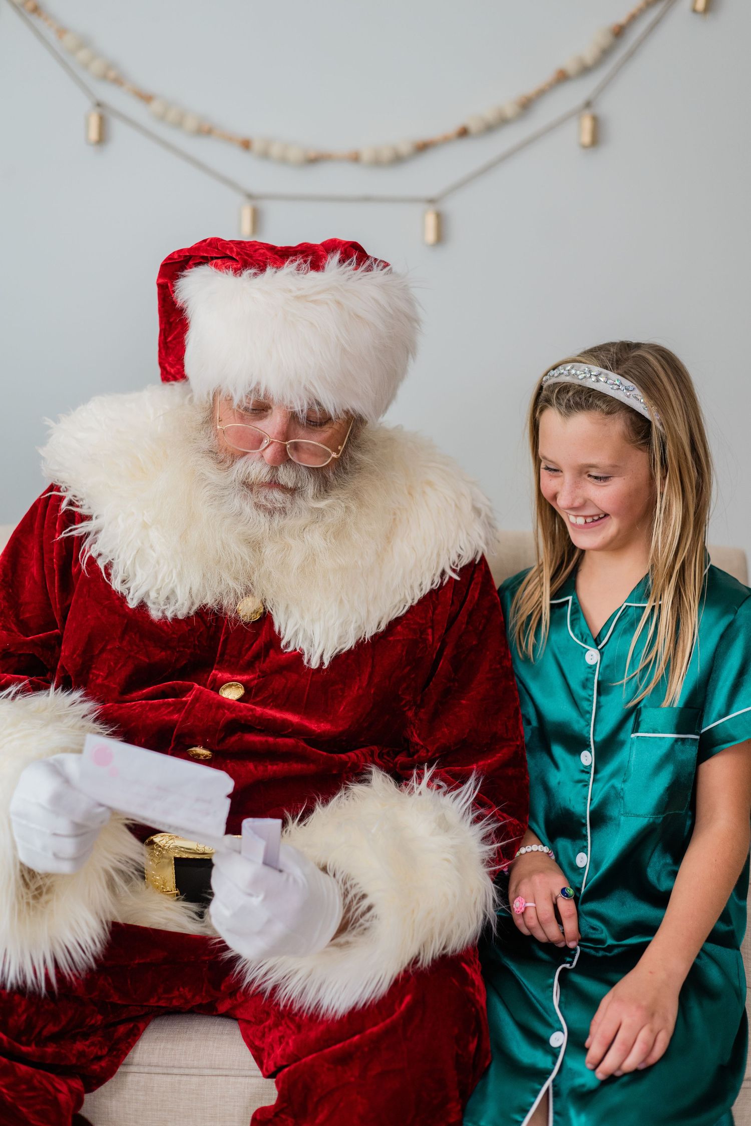 A young girl in green satin pajamas smiles while sitting with Santa Claus reading a letter indoors.
