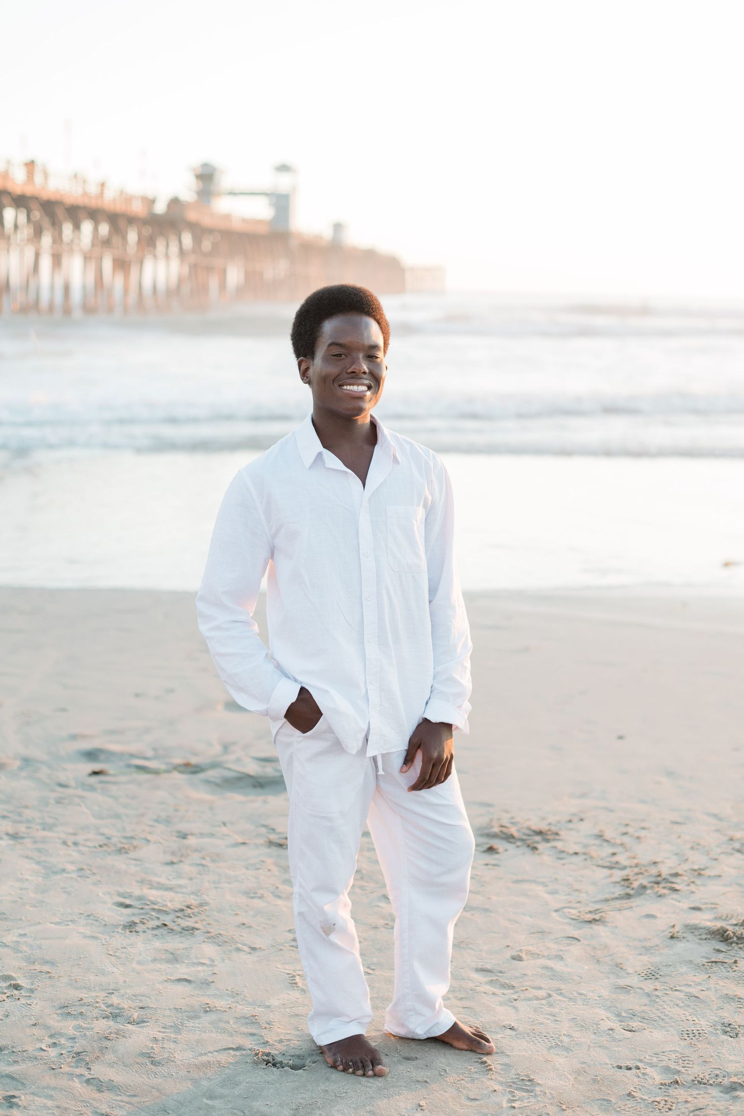 Smiling young Black man in all-white outfit standing barefoot on sandy beach with pier in background.