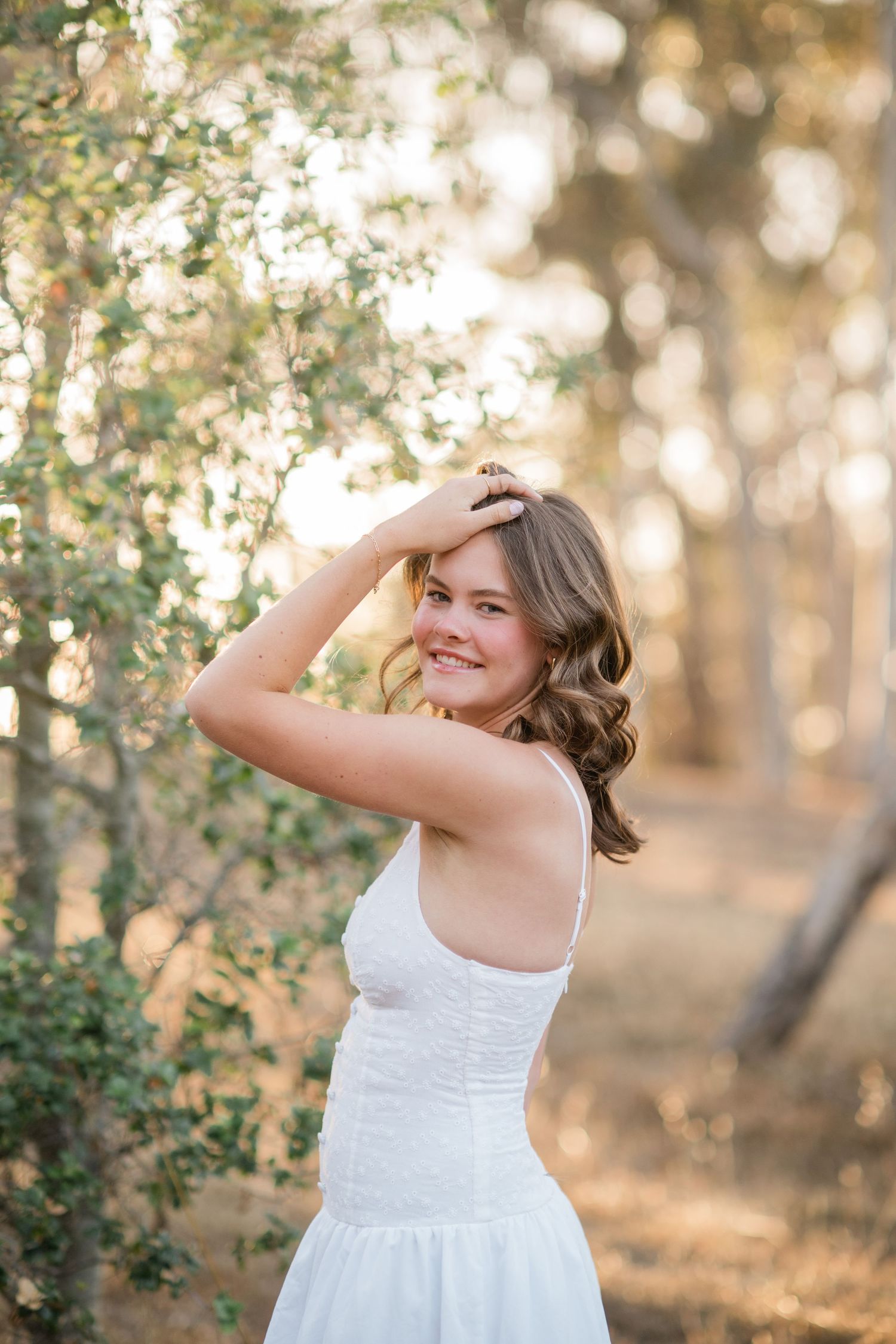 Young woman in white dress smiling outdoors among green trees in golden sunlight.