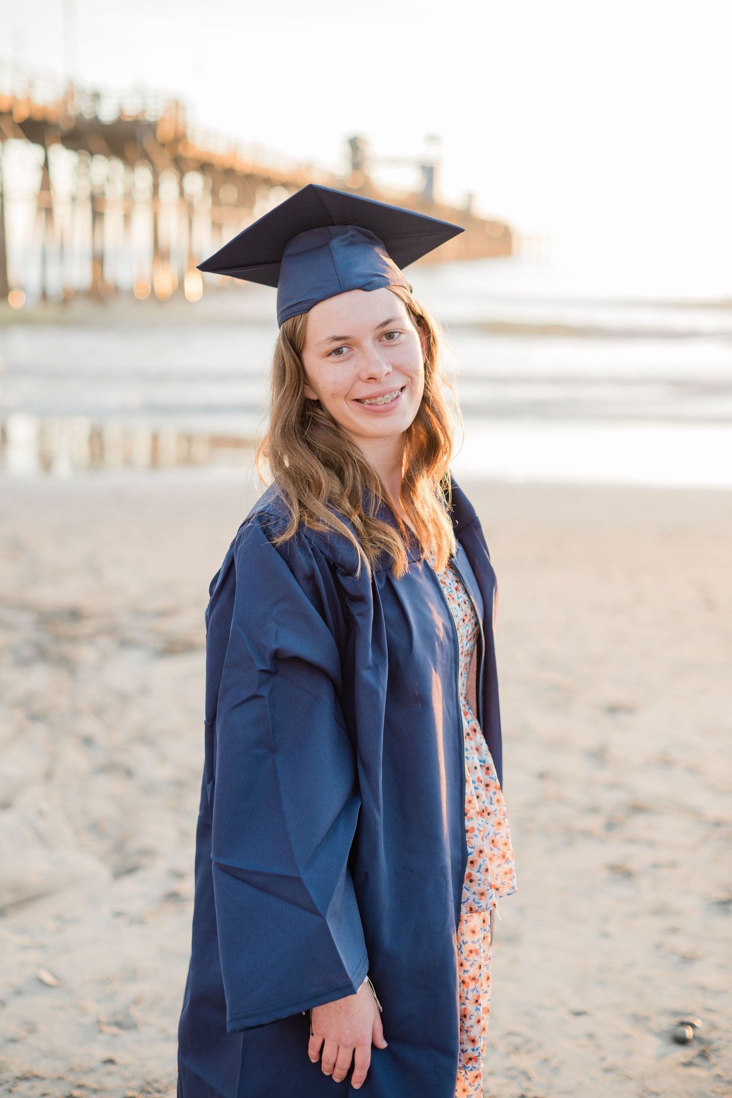 Young woman in navy blue graduation cap and gown smiling on a sandy beach with a pier in the background.