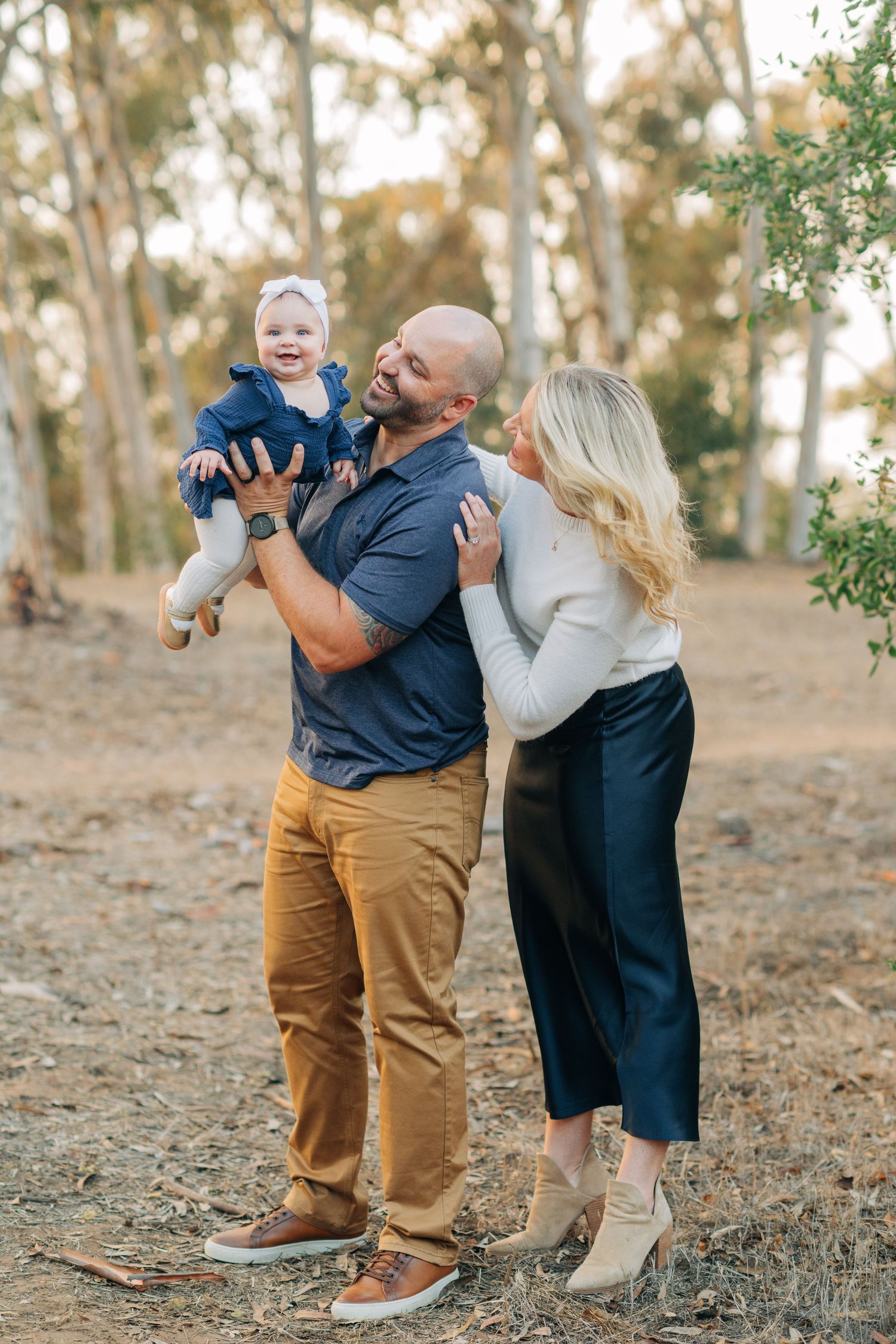 A happy family of three outdoors, father holding smiling baby girl while mother embraces him among eucalyptus trees.