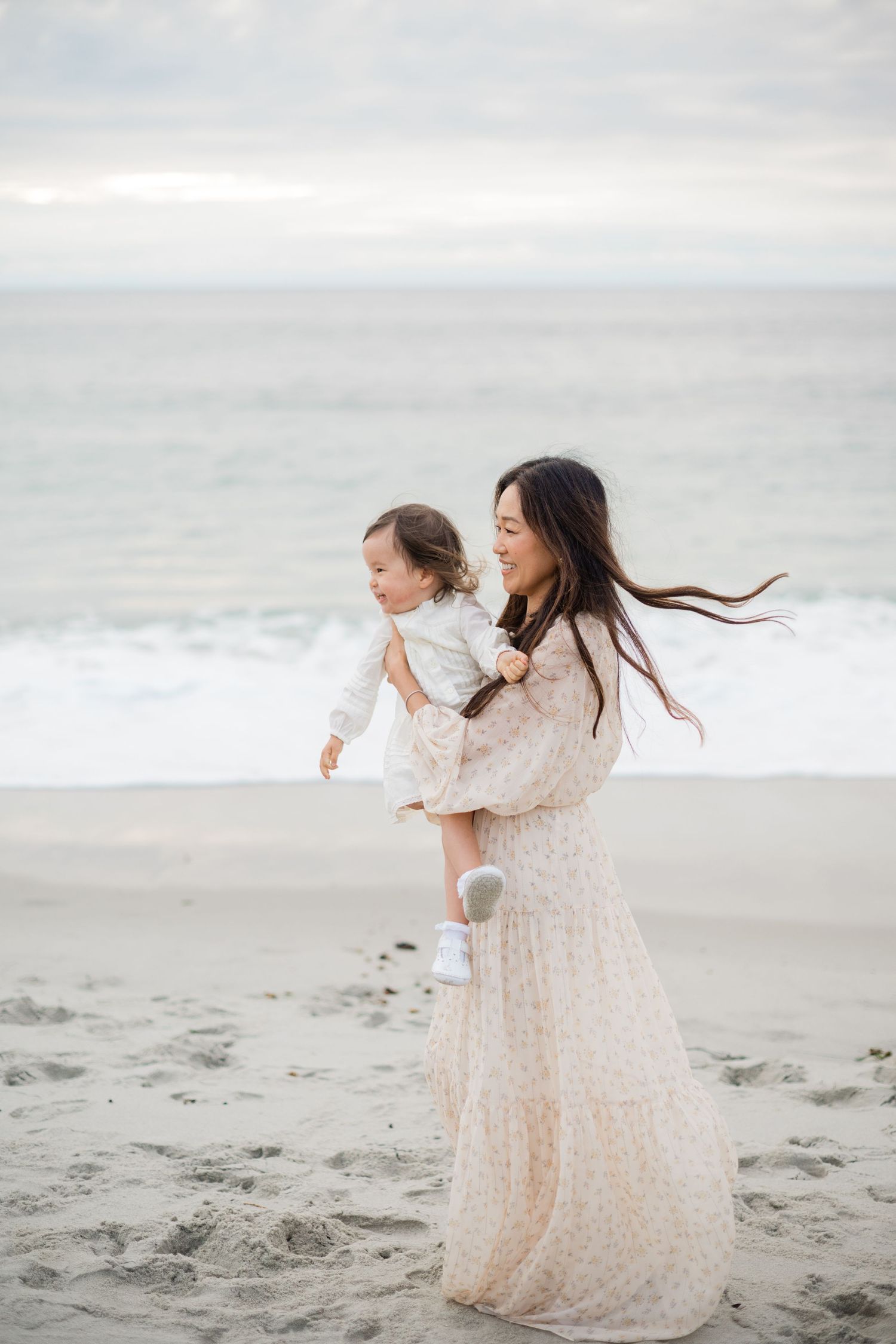 Mother in floral dress holding toddler girl on windy beach with ocean backdrop.