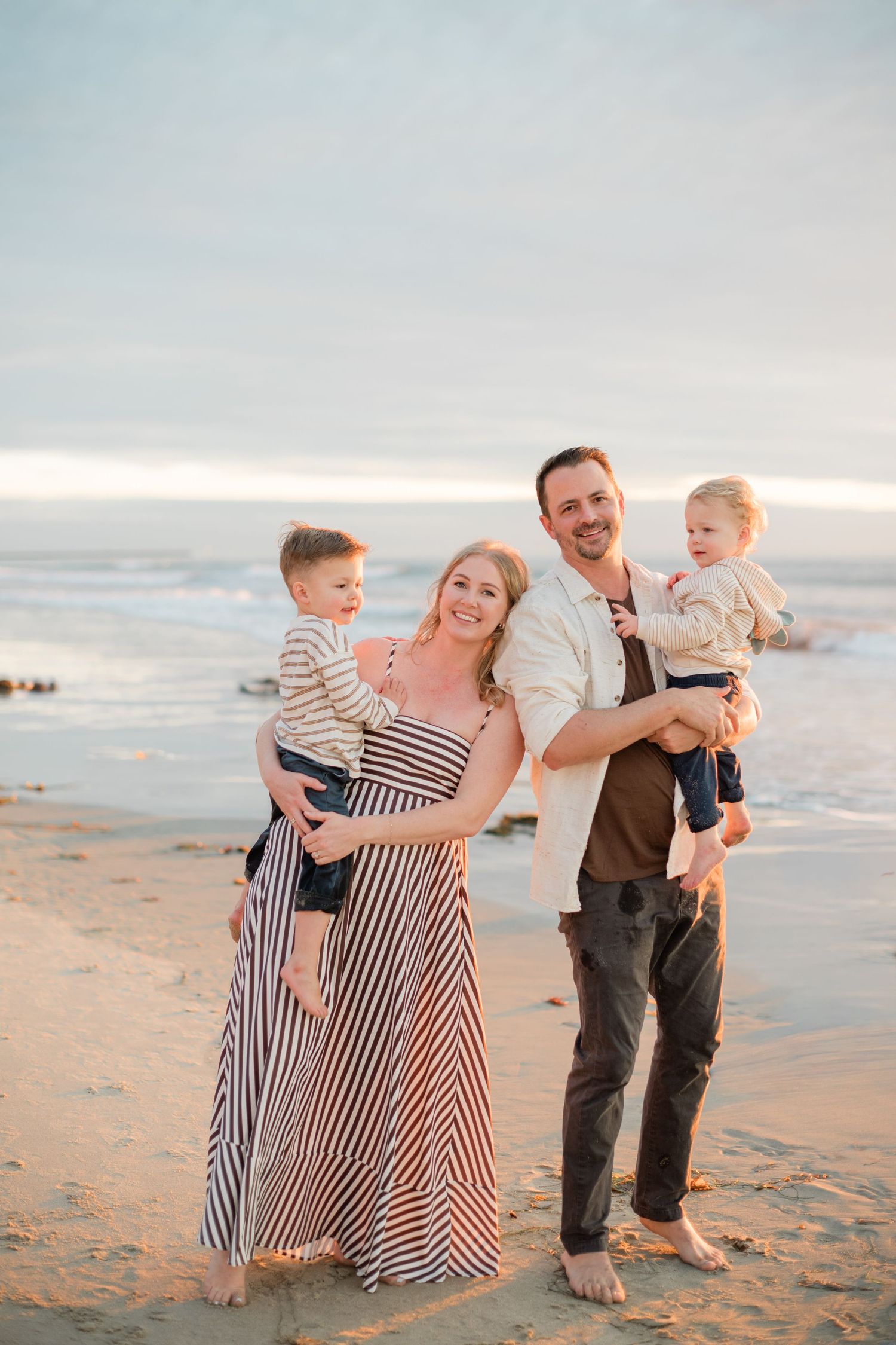 A happy family of four poses on a sandy beach at sunset, with parents holding two young toddler boys.
