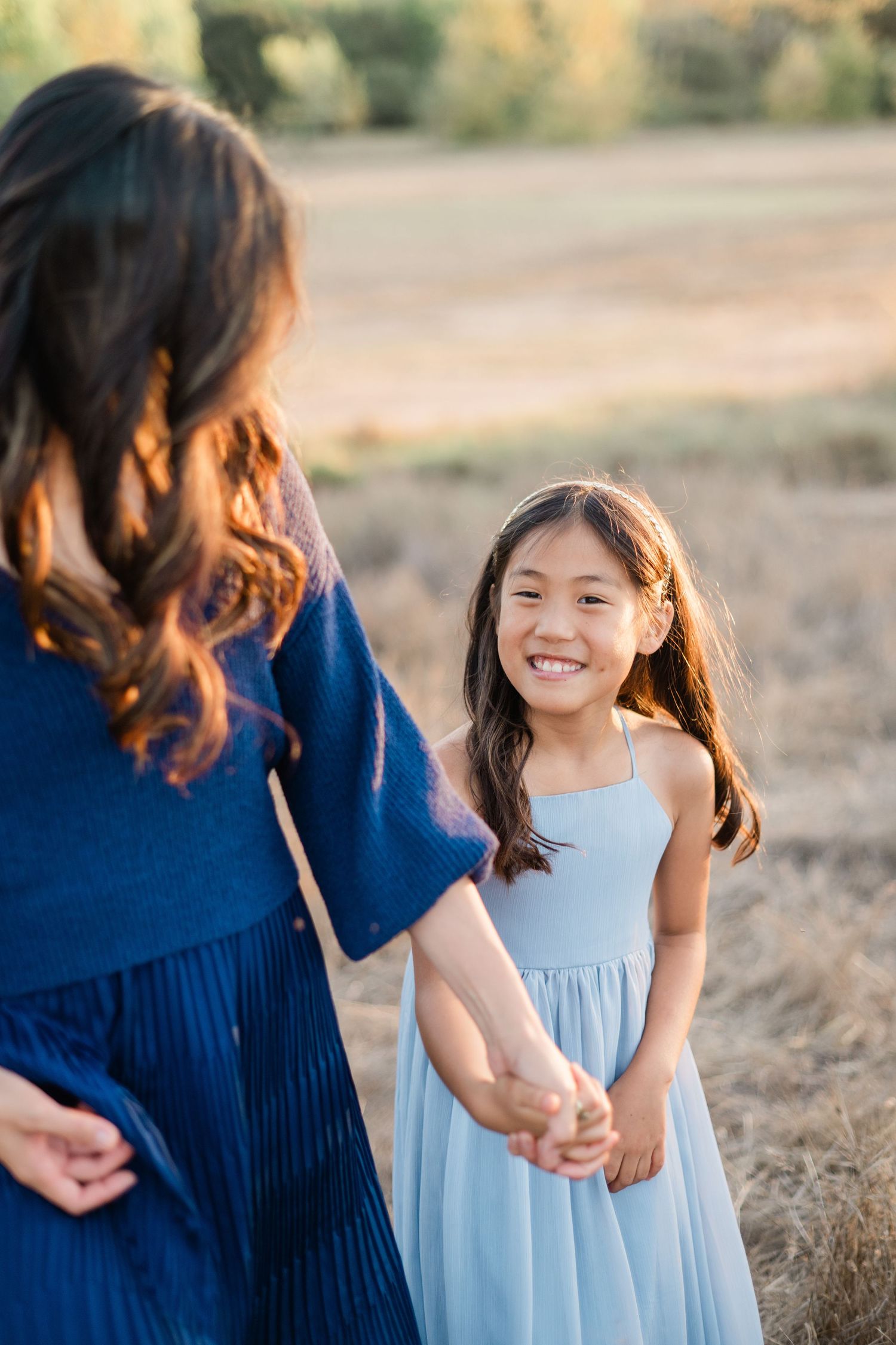 A smiling young Asian girl in a light blue dress holds hands with a woman in a navy dress in a golden field.