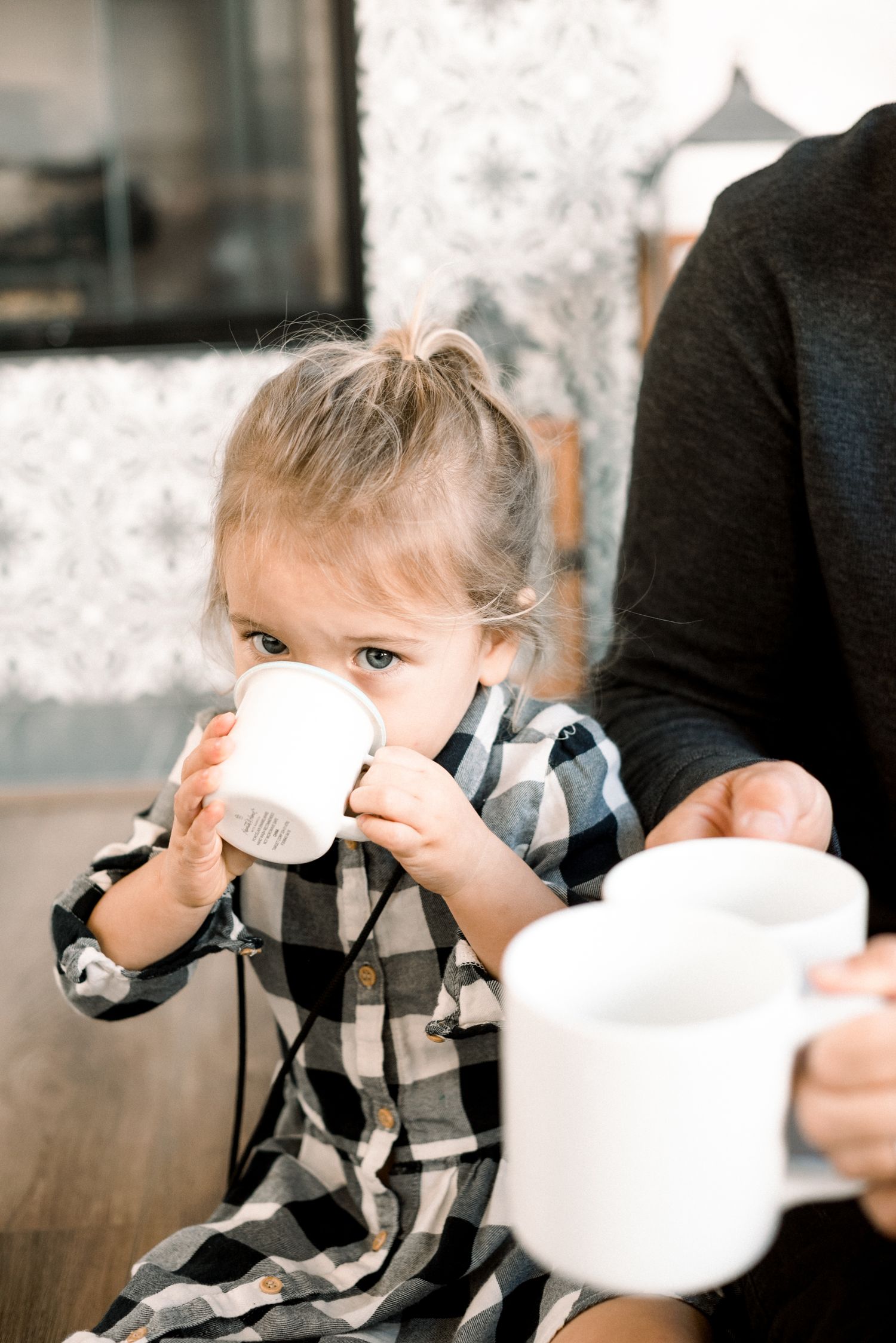 A toddler girl in a plaid dress drinks from a small white cup beside an adult holding a mug.