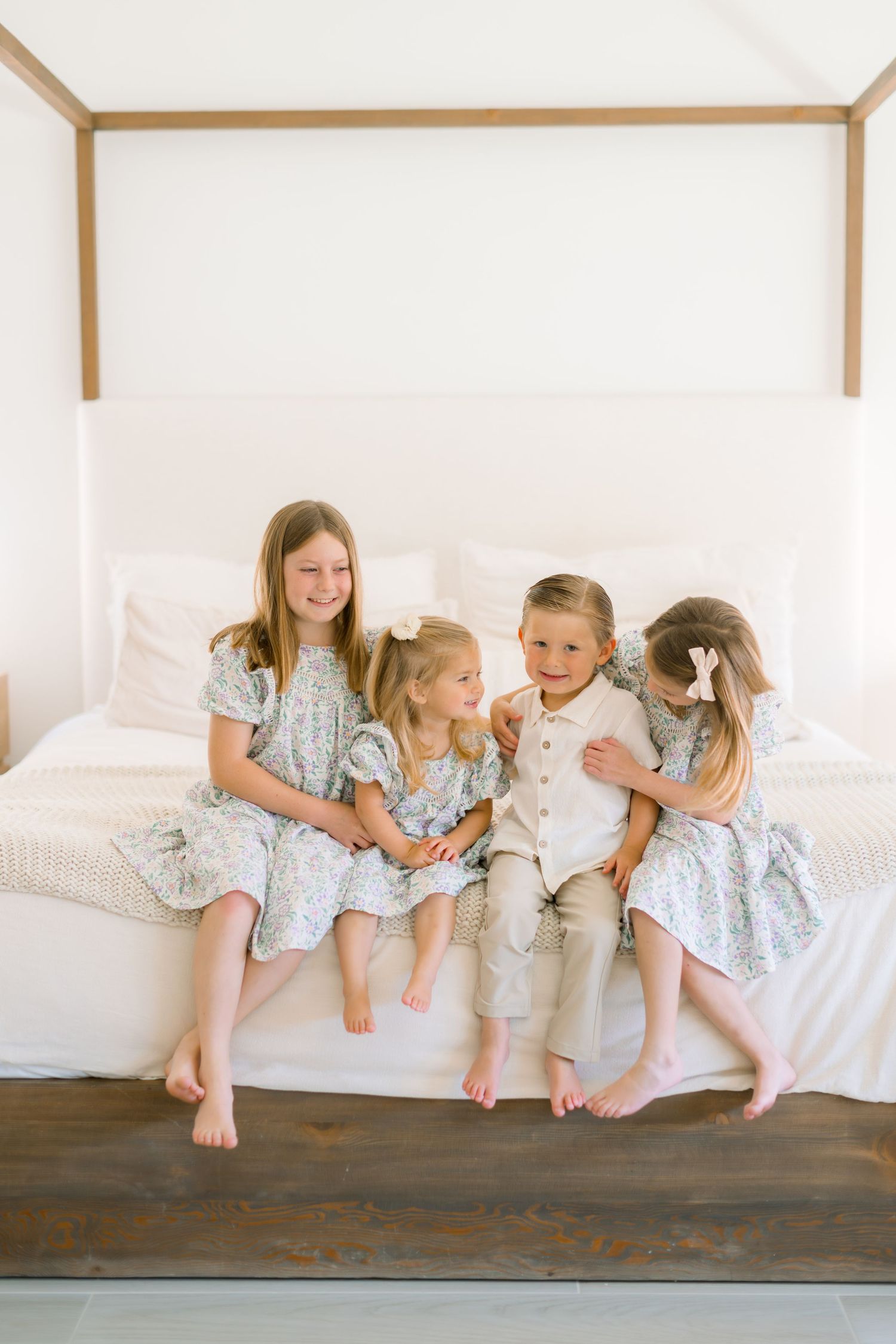 Four young children in floral and neutral outfits sit together smiling on a white bed in a bright bedroom.