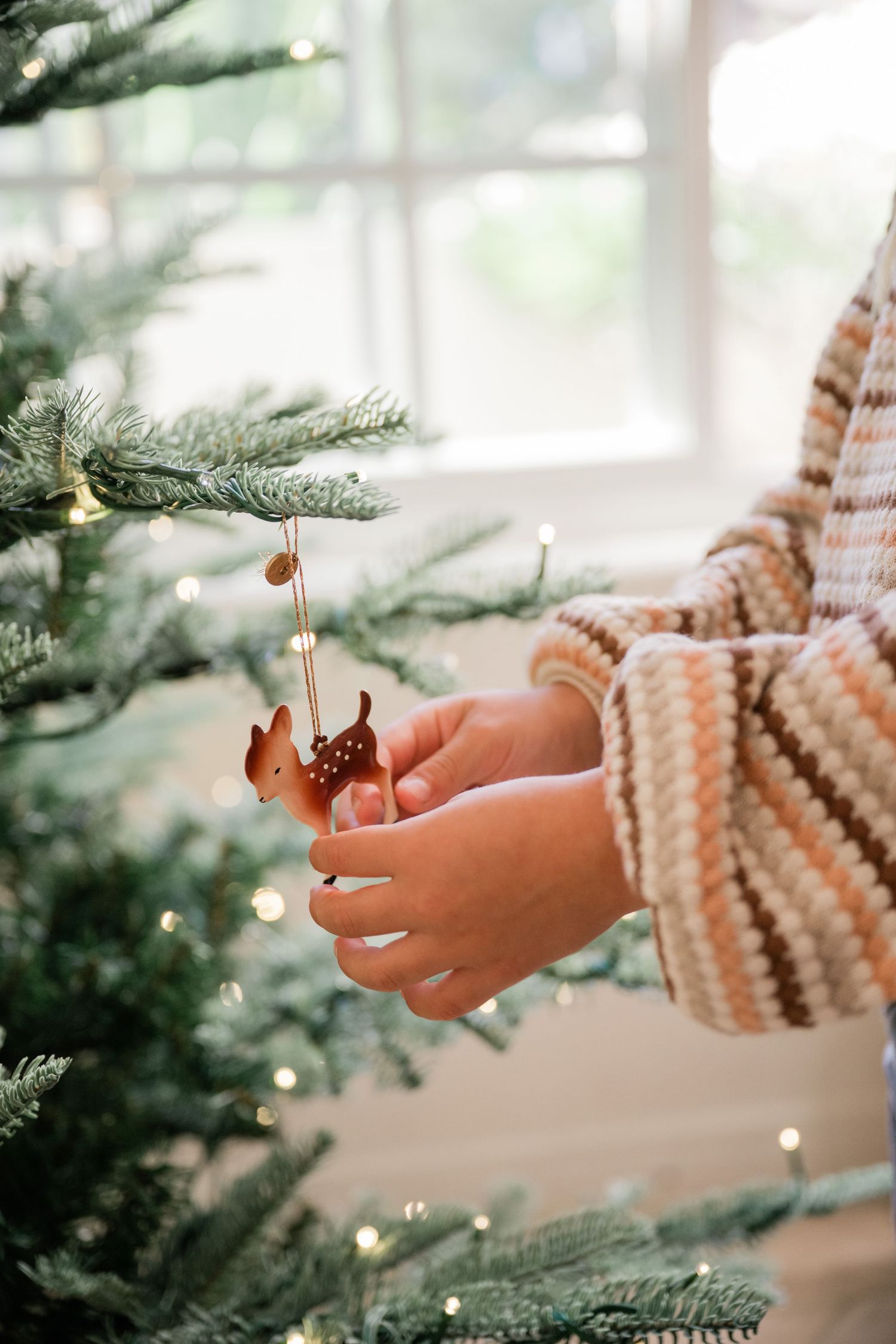 Child in striped sweater hanging a deer ornament on a Christmas tree.