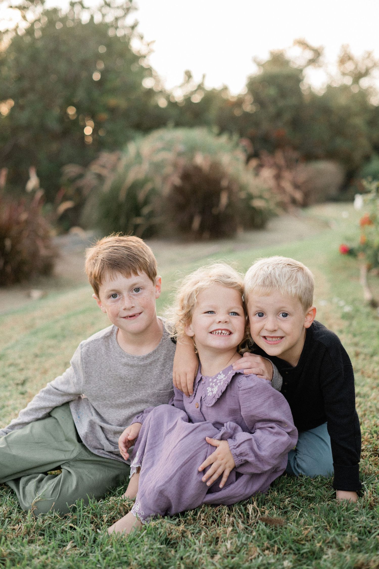 Three young siblings smile and cuddle together on green grass during a golden hour outdoor photo session.