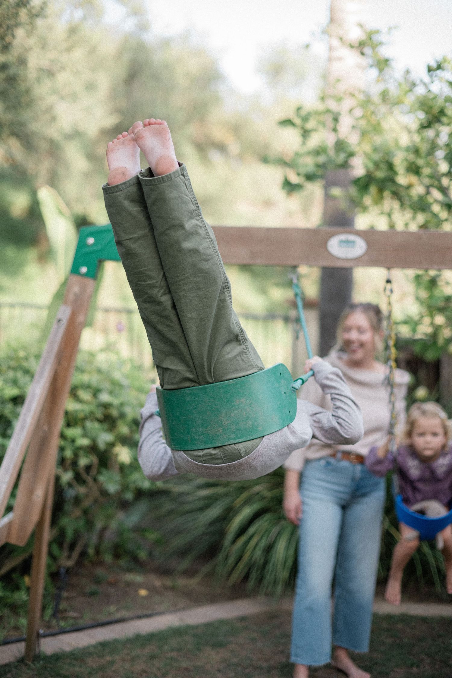 Child flipping upside down on a green tire swing in a backyard playground while a woman watches nearby.