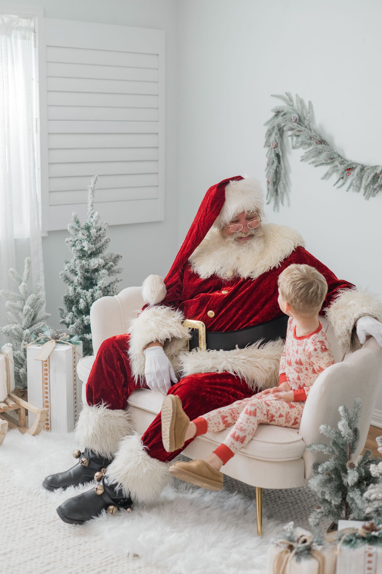 Santa Claus reads to a young child in pajamas while seated in a white armchair amid Christmas decor.