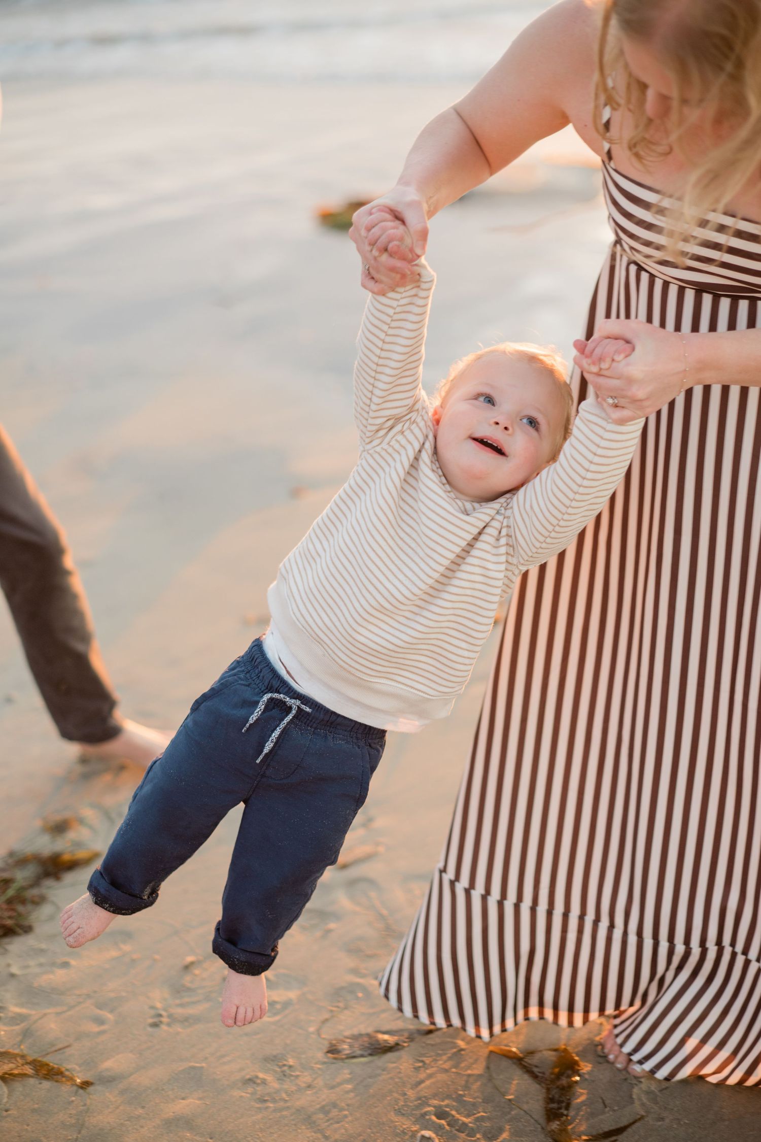 Joyful toddler lifted by mother on sandy beach at sunset wearing striped shirt and jeans.