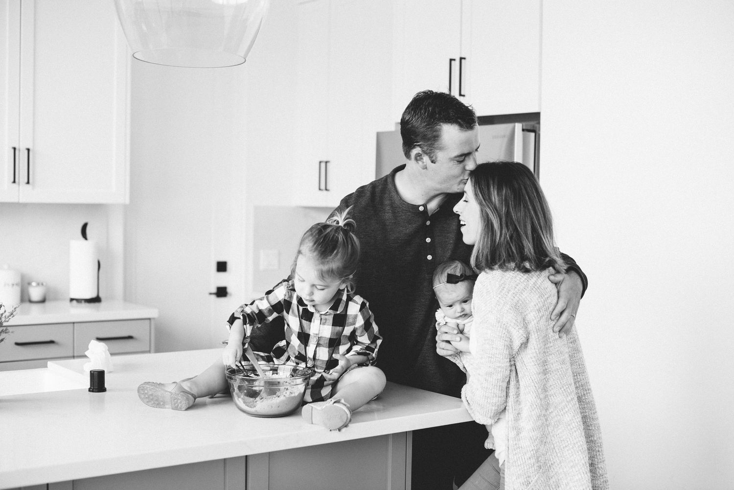 Black and white photo of a family cooking together in a modern kitchen, parents and two young children at the counter.