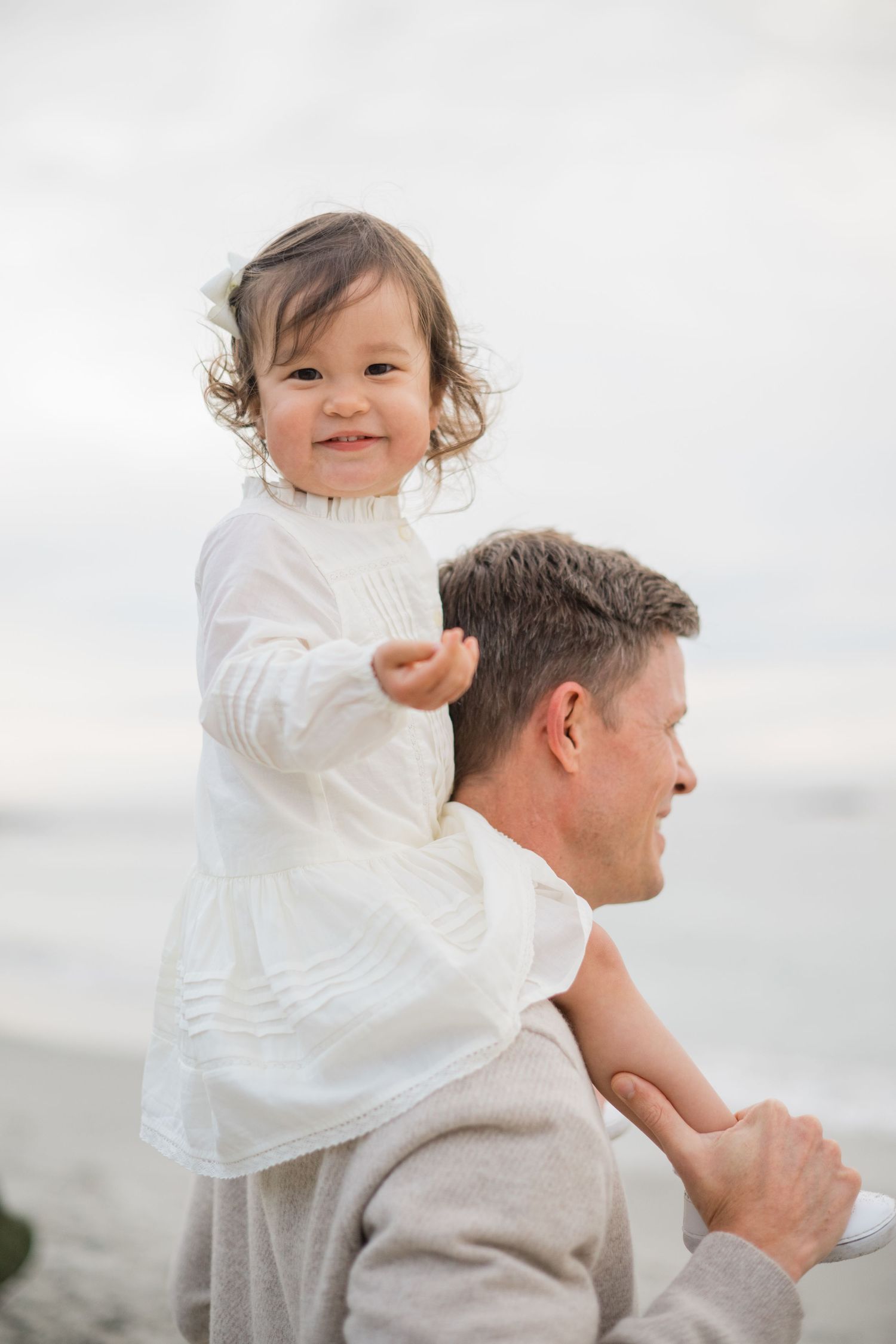 Smiling toddler girl in white dress sitting on fathers shoulders at the beach.