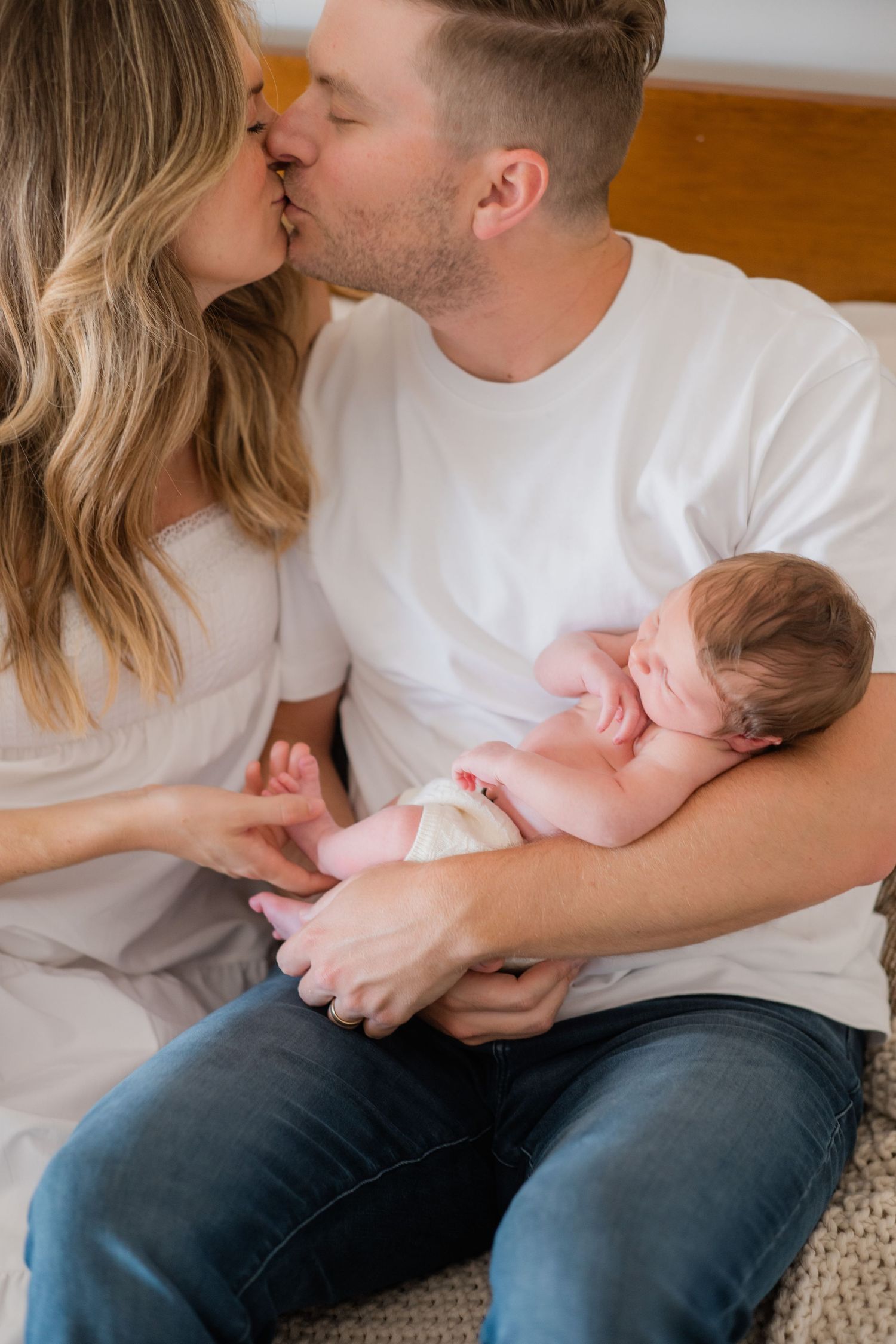 A couple shares a tender kiss while cradling their newborn baby together in a warm family portrait.