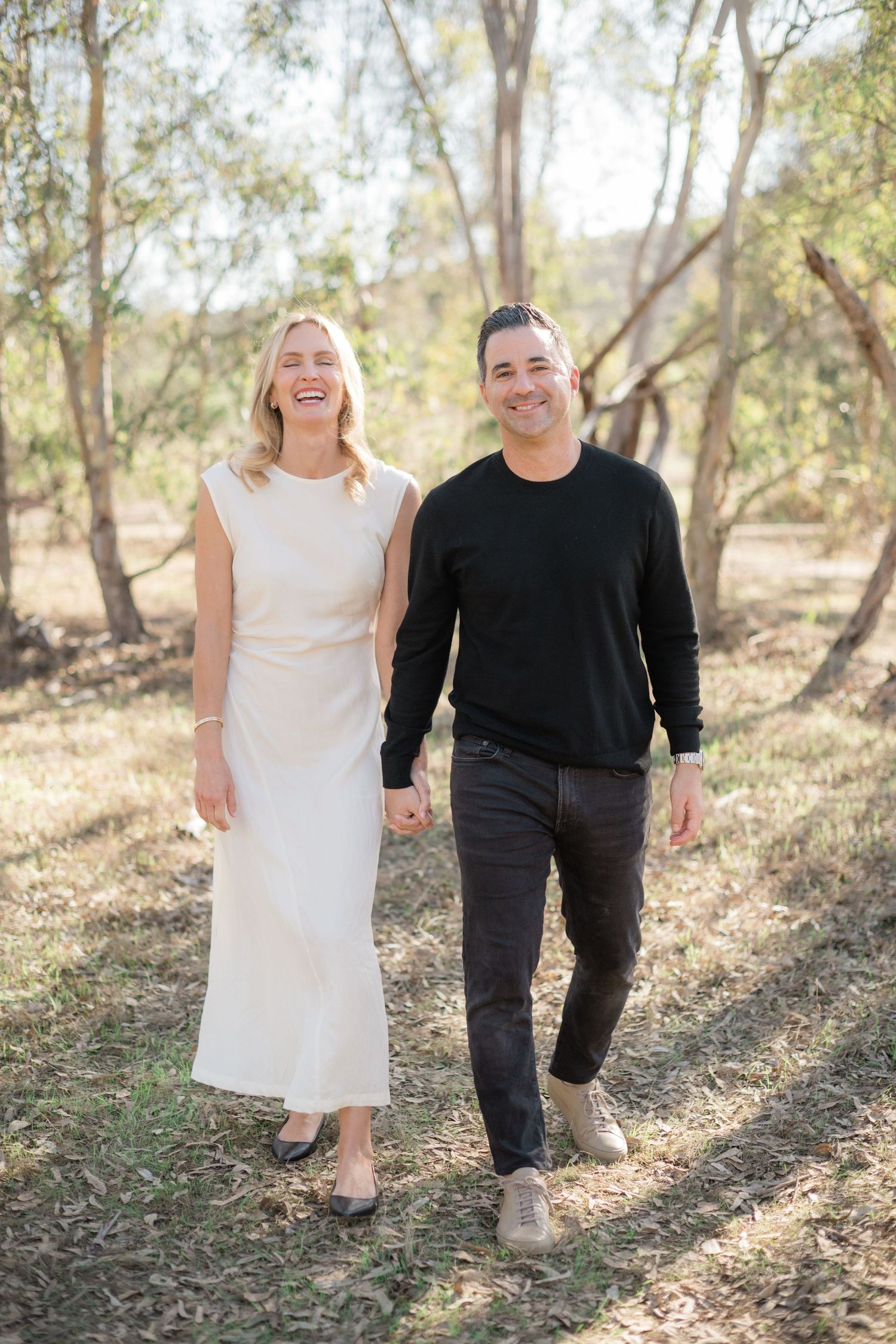 A smiling couple walks hand-in-hand outdoors among trees, woman in white dress, man in black sweater and jeans.