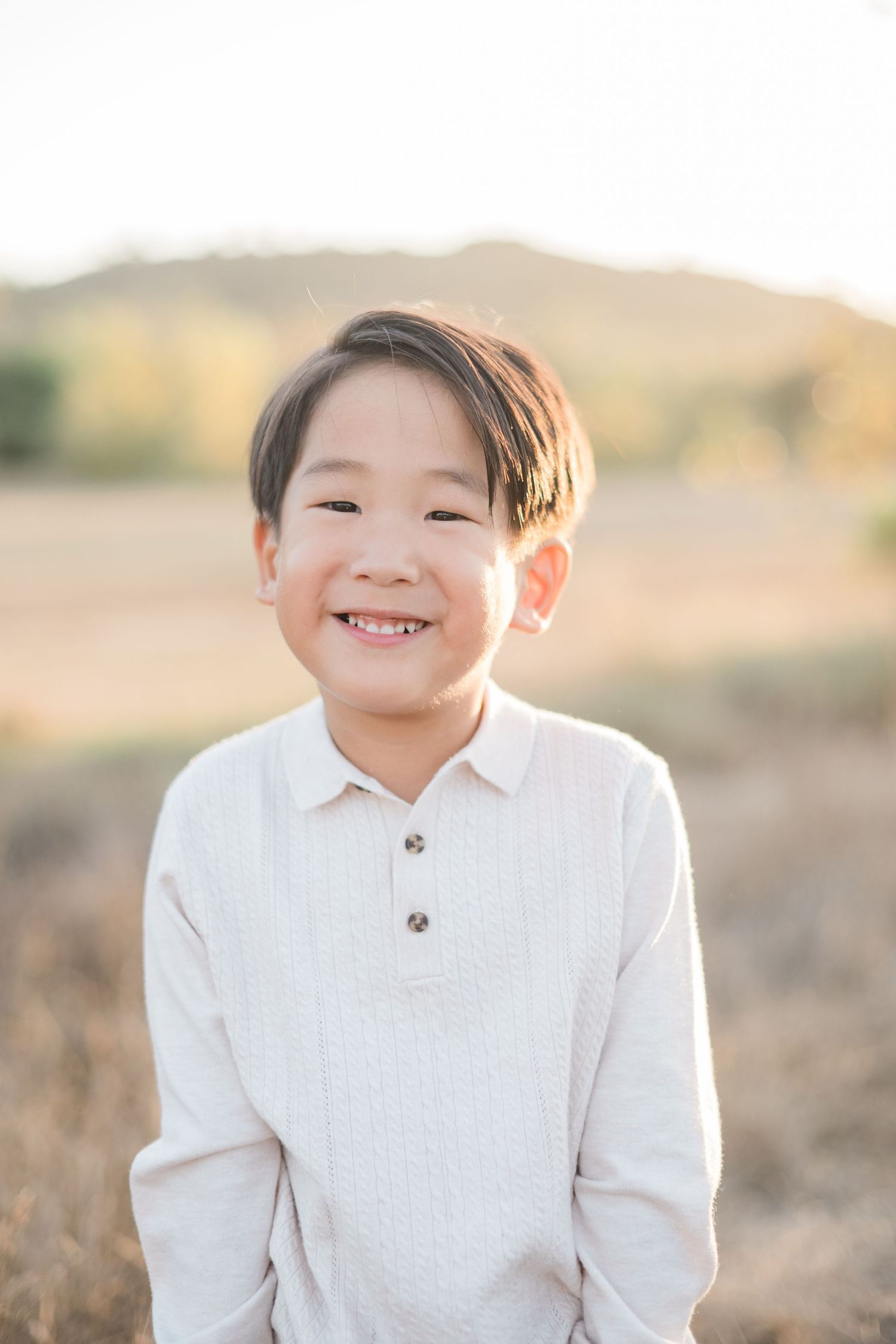 Smiling young Asian boy in white shirt standing in golden field with hills in background.