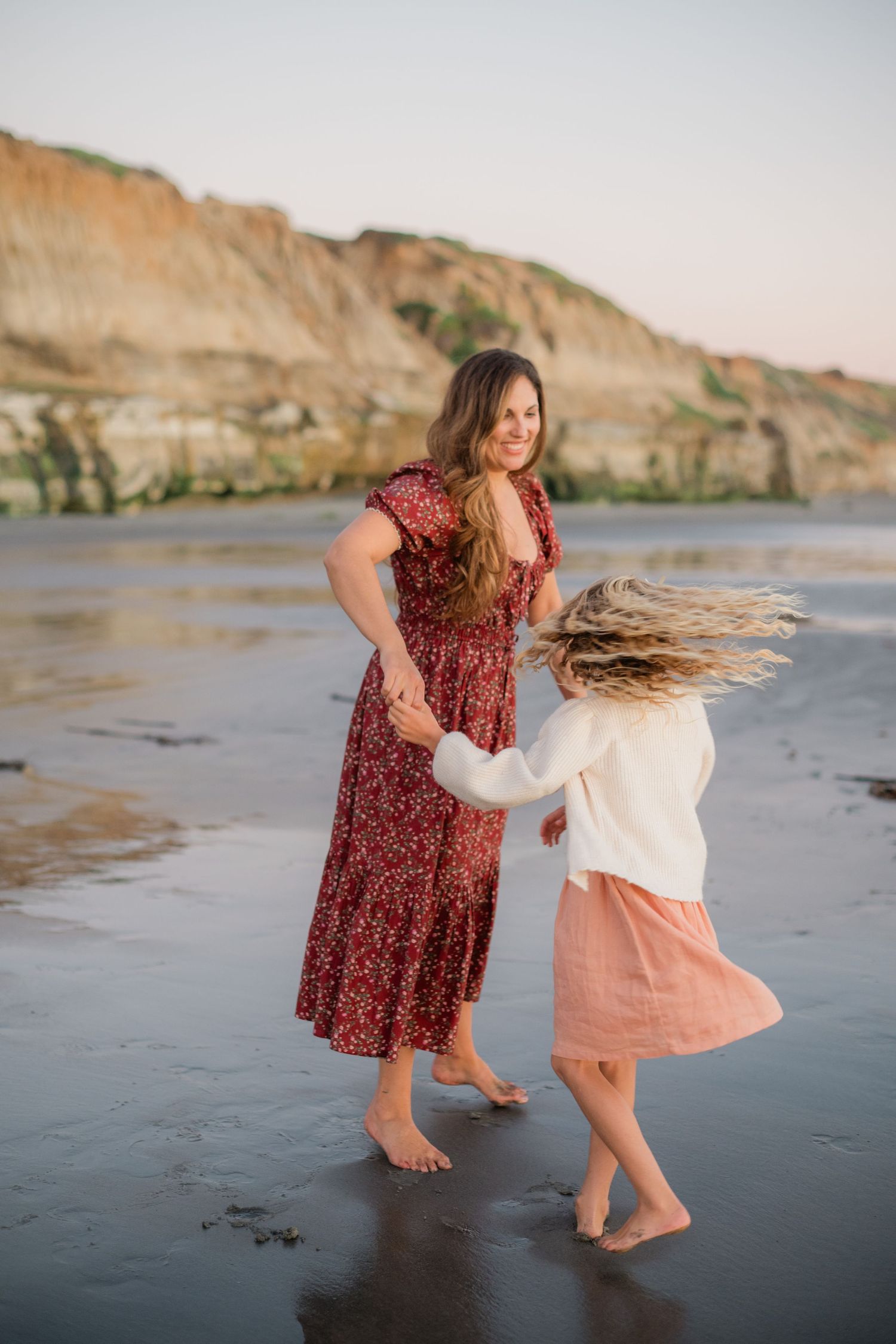 A mother and daughter dance barefoot on a sandy beach at sunset, with coastal cliffs in the background.