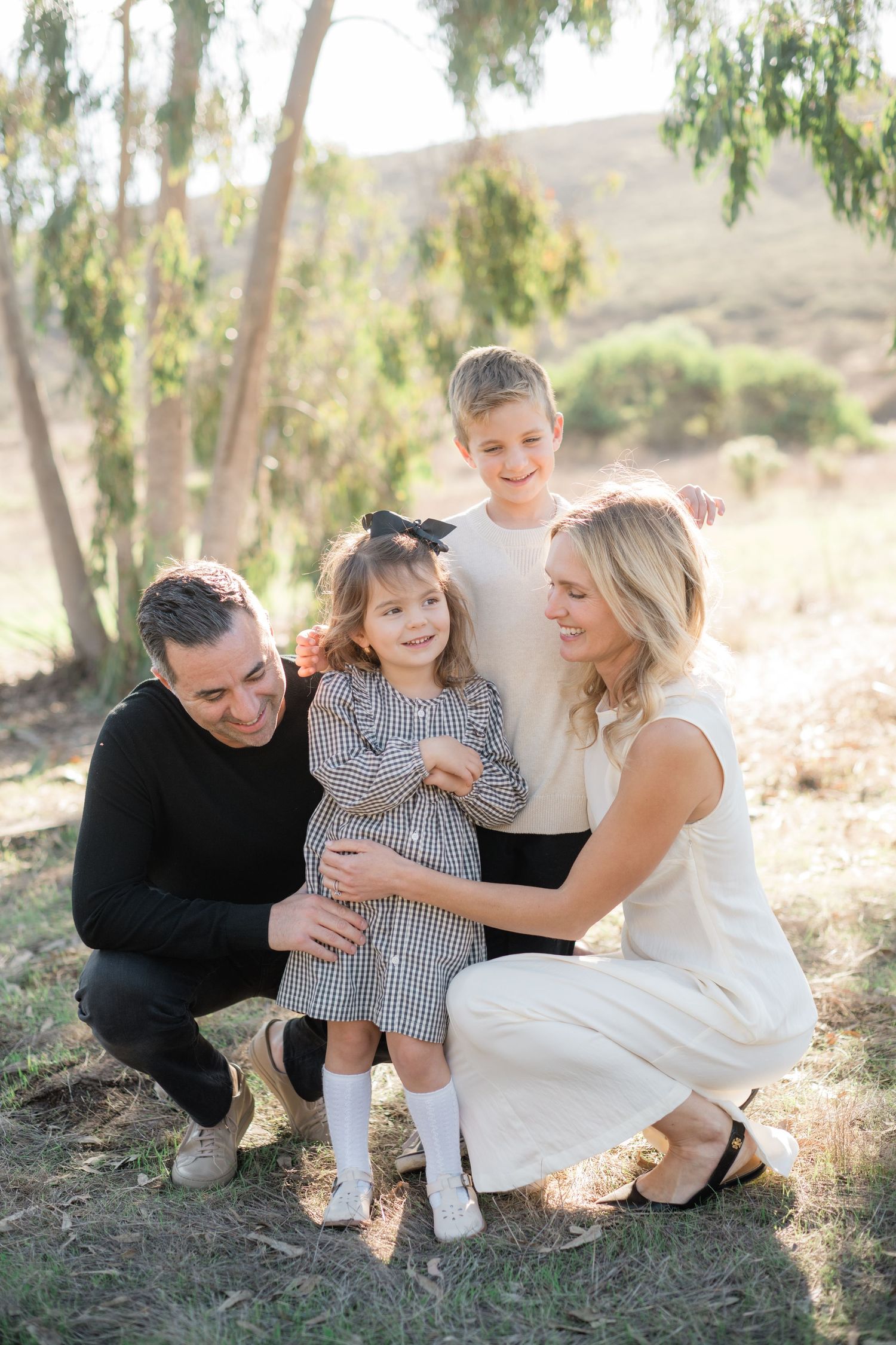 A happy family of four poses outdoors among trees, with parents crouching beside their young daughter and son.