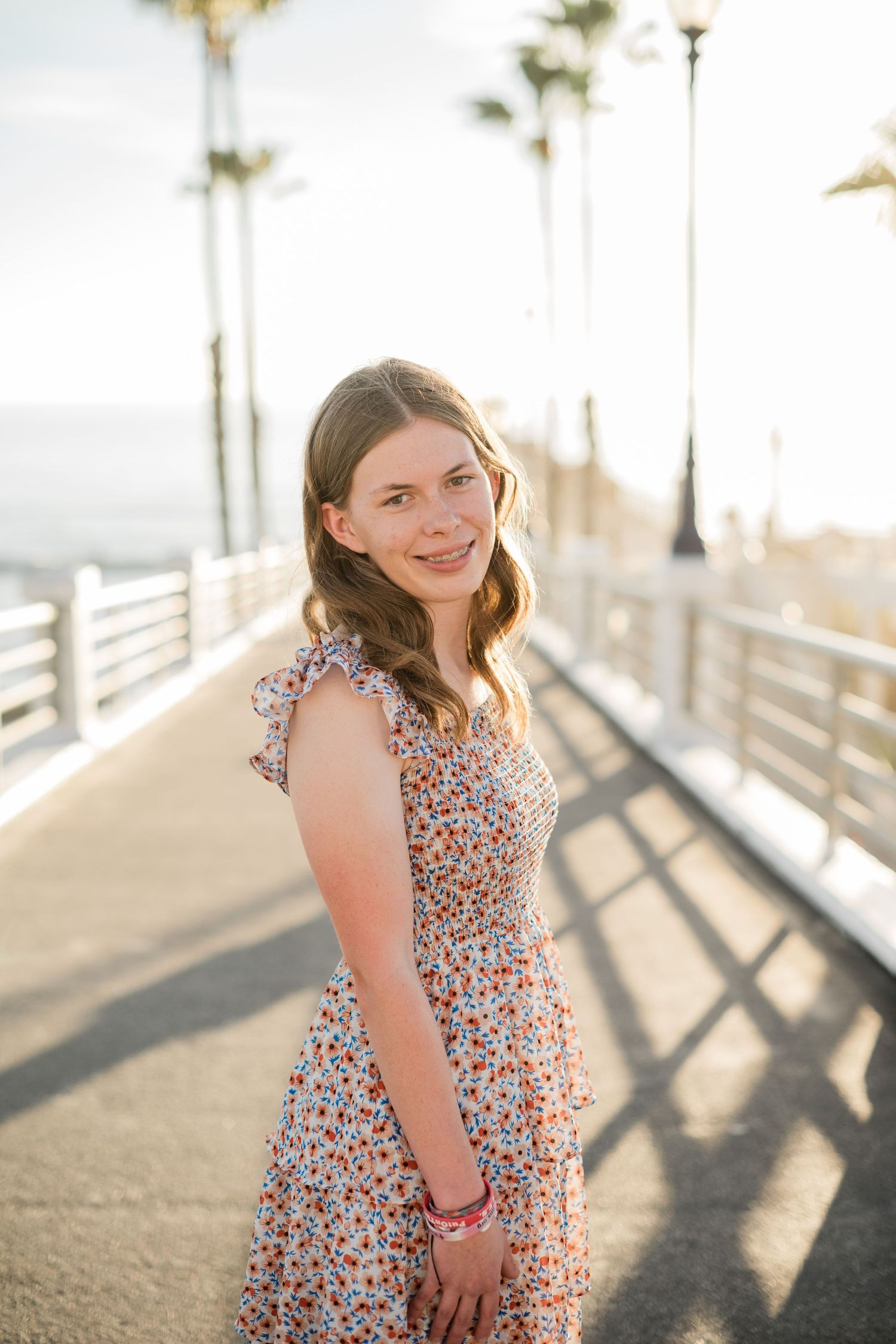 Young woman in floral dress smiling on a sunny pier with palm trees in the background.