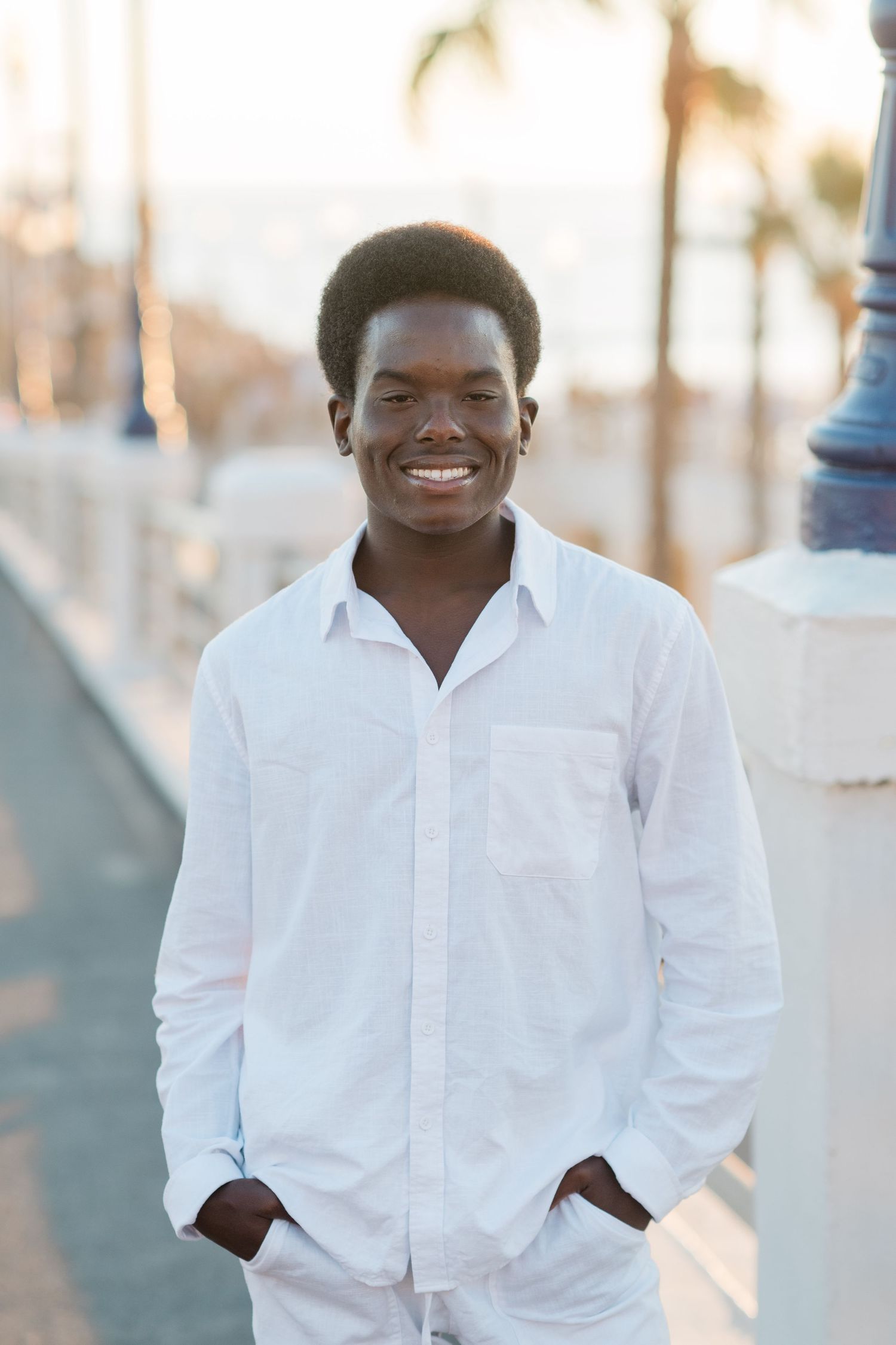 Smiling young Black man in white shirt standing on sunny boardwalk with palm trees in background.