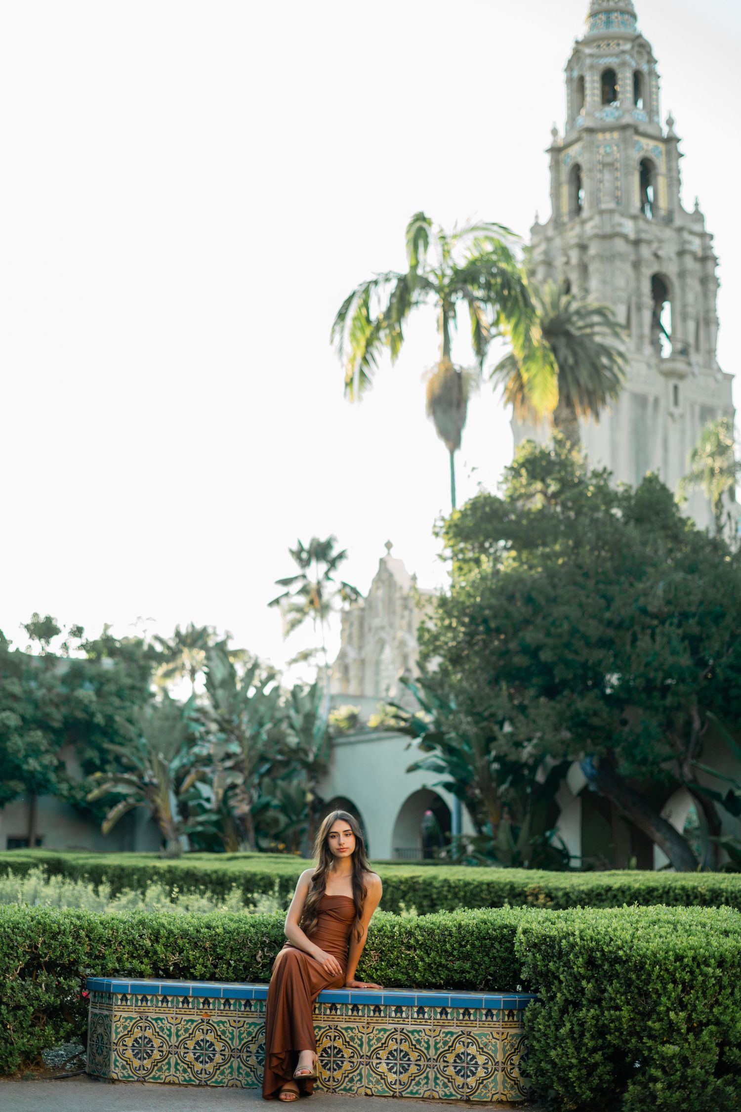 Woman in brown outfit sitting on a decorative tiled bench at Balboa Park with California Tower in background.
