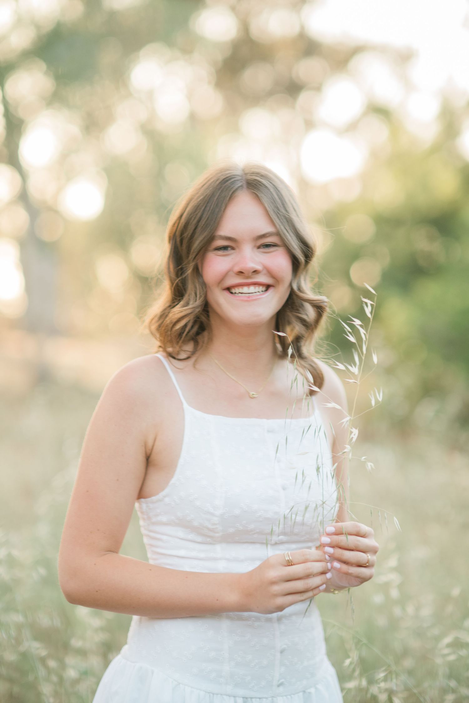 Smiling young woman in white sundress holding wildgrass in a sunlit field with bokeh background.