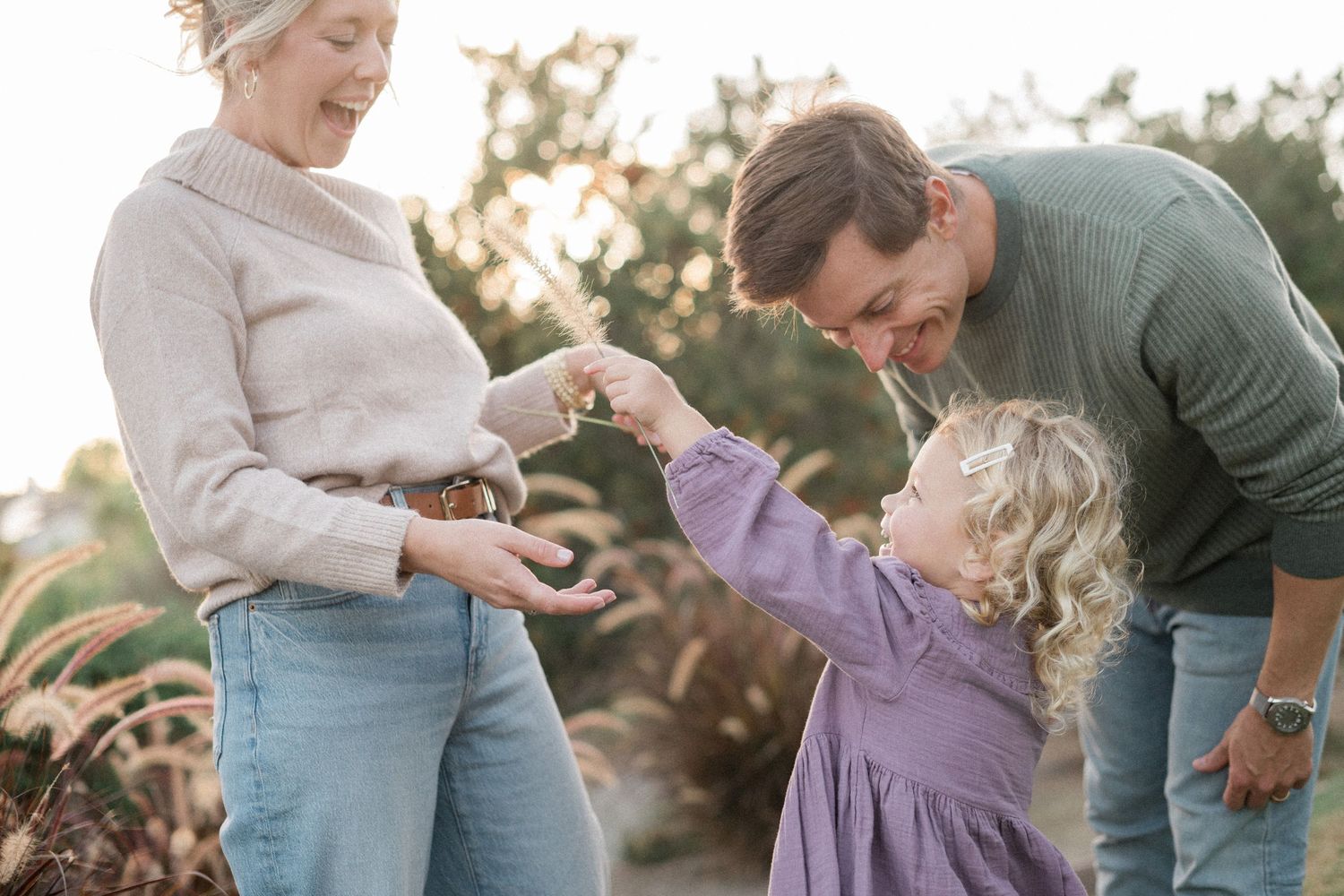 A joyful family of three shares a tender moment outdoors, with parents engaging their curly-haired toddler girl in purple dress.