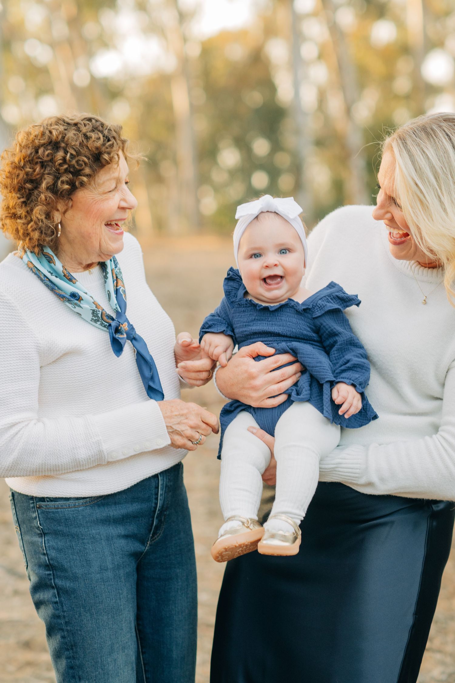 Two women in white sweaters smiling at a happy baby girl in a blue dress outdoors in autumn.
