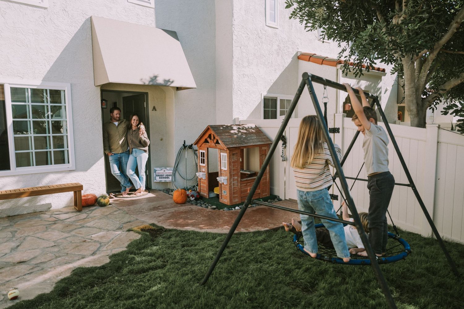 Children playing in backyard with swing set and playhouse while parents watch from doorway.