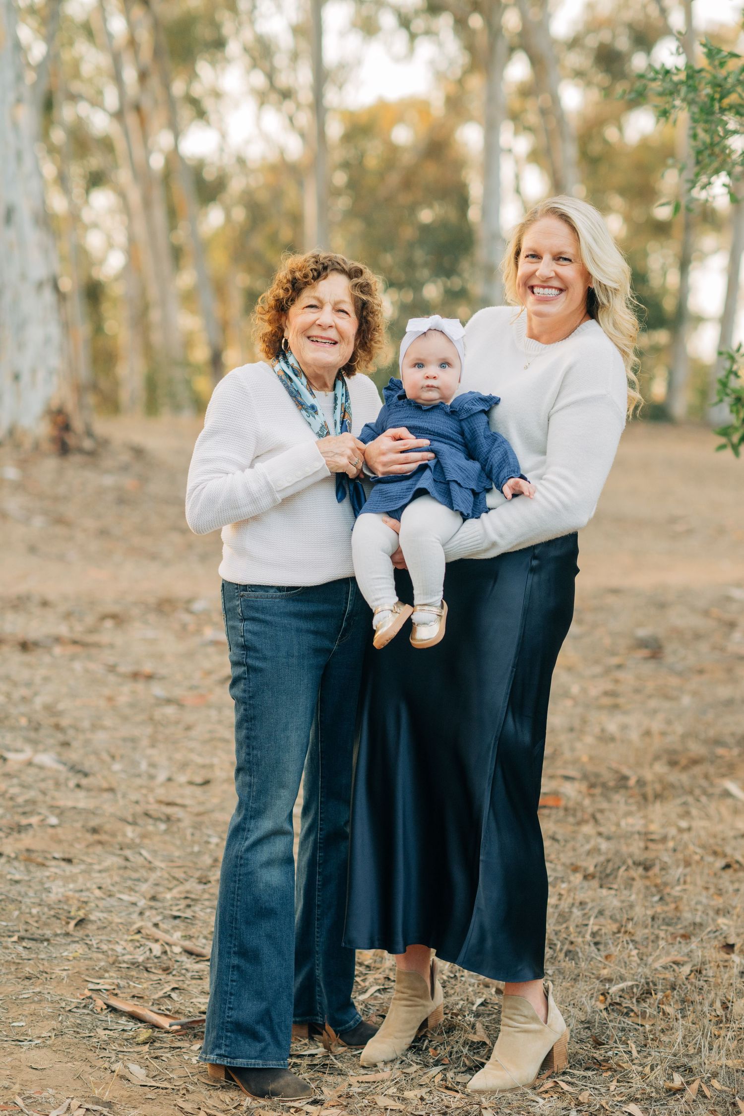 Three generations of women pose outdoors among eucalyptus trees, with grandmother, mother, and baby girl in matching white and blue outfits.