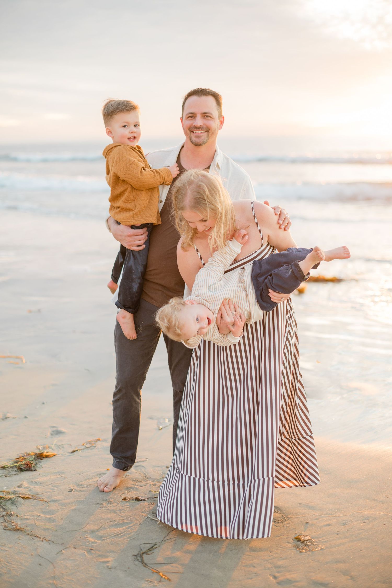 Happy family of five posing together on a sandy beach at sunset, parents holding three young children.