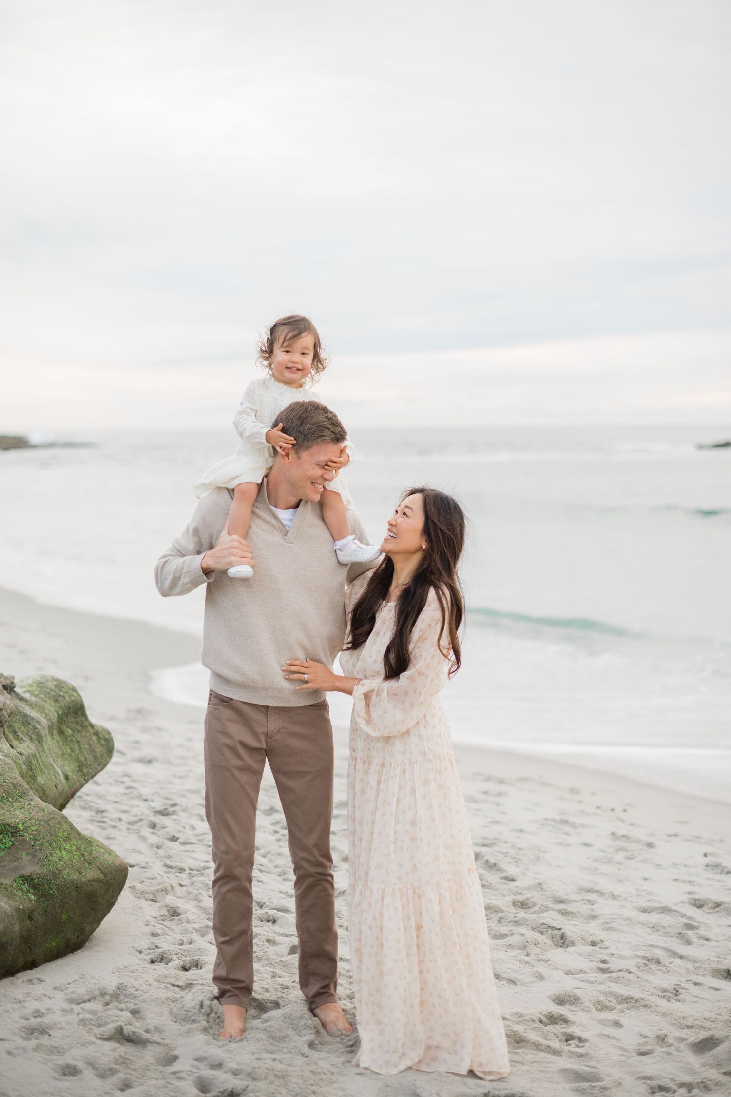 A happy family of three poses on a sandy beach, with dad holding toddler on shoulders beside smiling mom.
