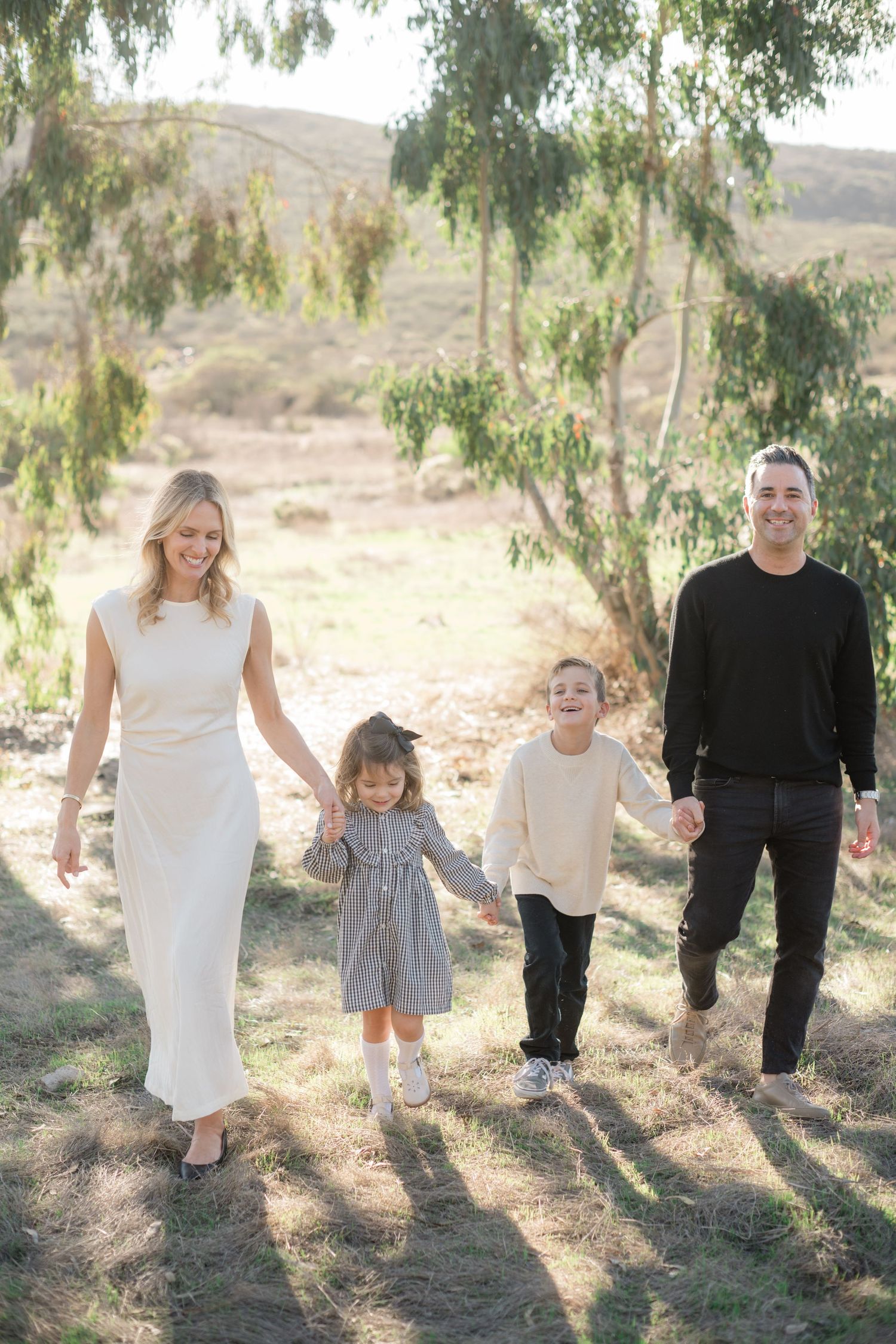 Joyful family of four holding hands while walking outdoors among eucalyptus trees in natural light.