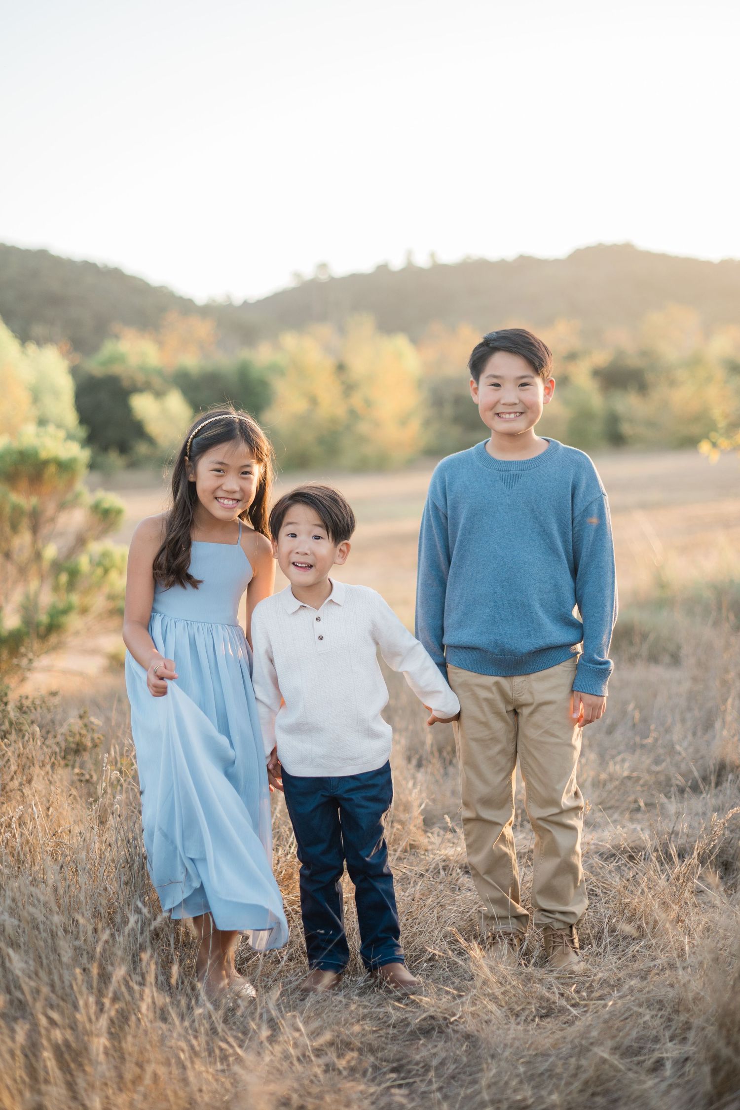 Three smiling Asian children pose together in a golden field at sunset, wearing coordinating blue and tan outfits.