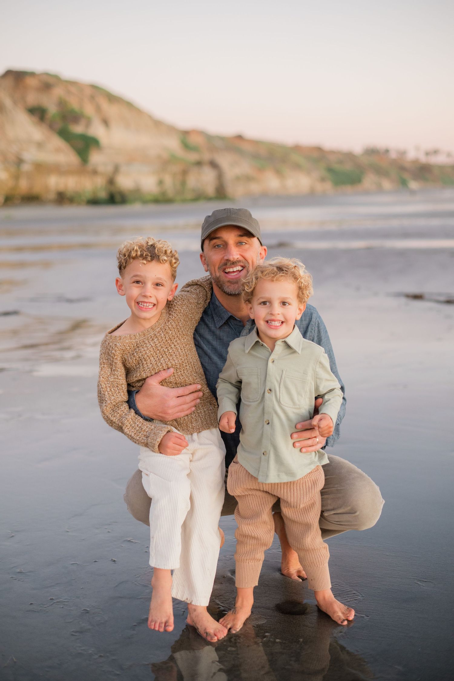 Smiling father in cap crouches with his two young sons on a sandy beach at sunset.