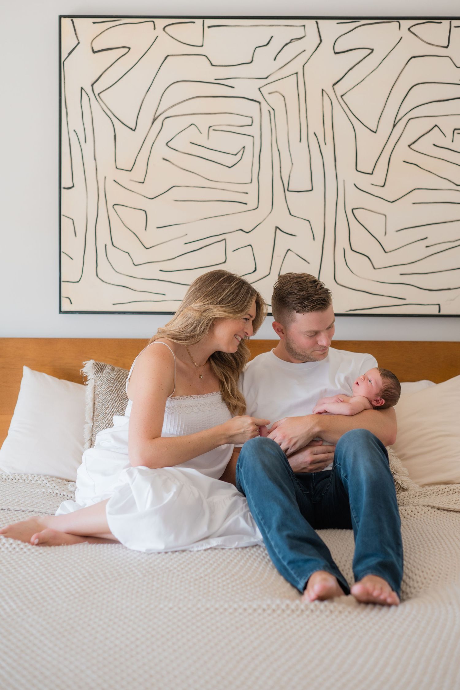 A couple sits on a bed admiring their newborn baby, with a large abstract line art painting on the wall behind them.