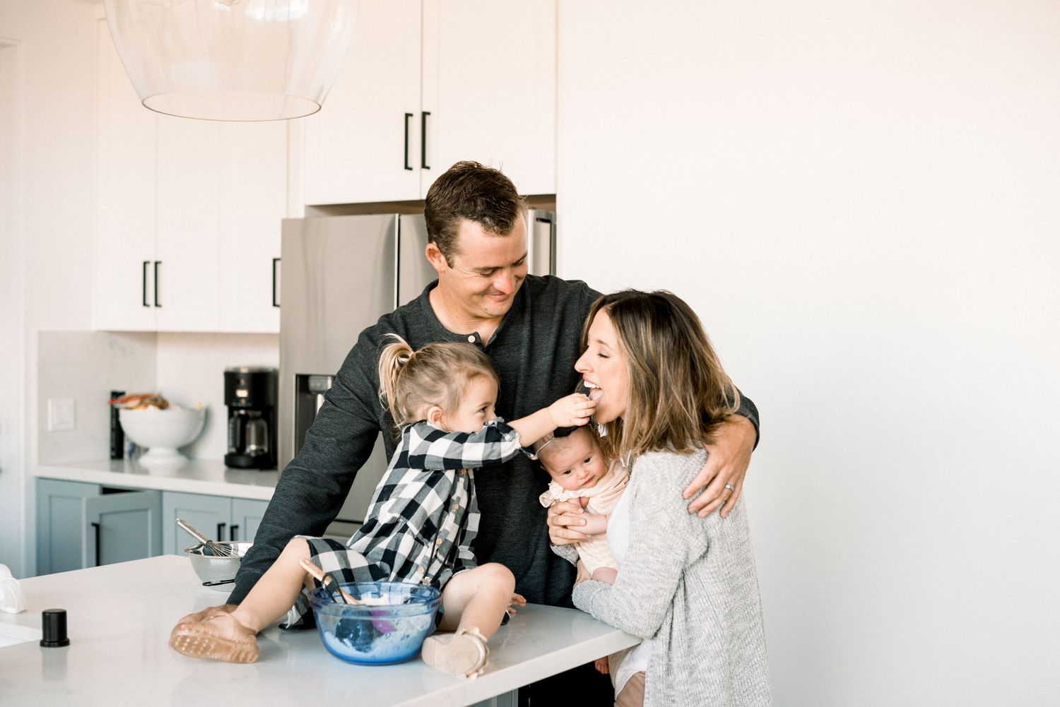 Family of four sharing a joyful moment in a bright modern kitchen, with dad holding baby and young daughter nearby.