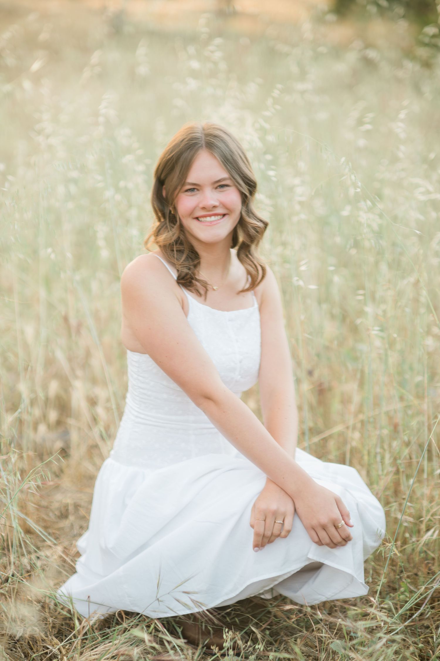 Smiling young woman in white dress sitting in golden meadow grass for outdoor portrait photo.