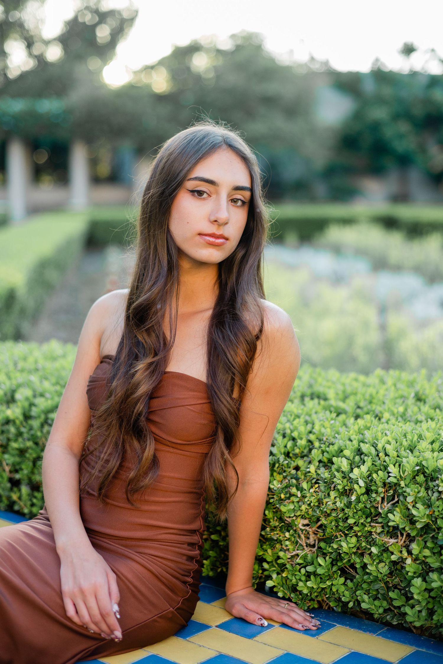 Young woman in brown strapless dress sitting beside manicured garden hedges in golden hour light.