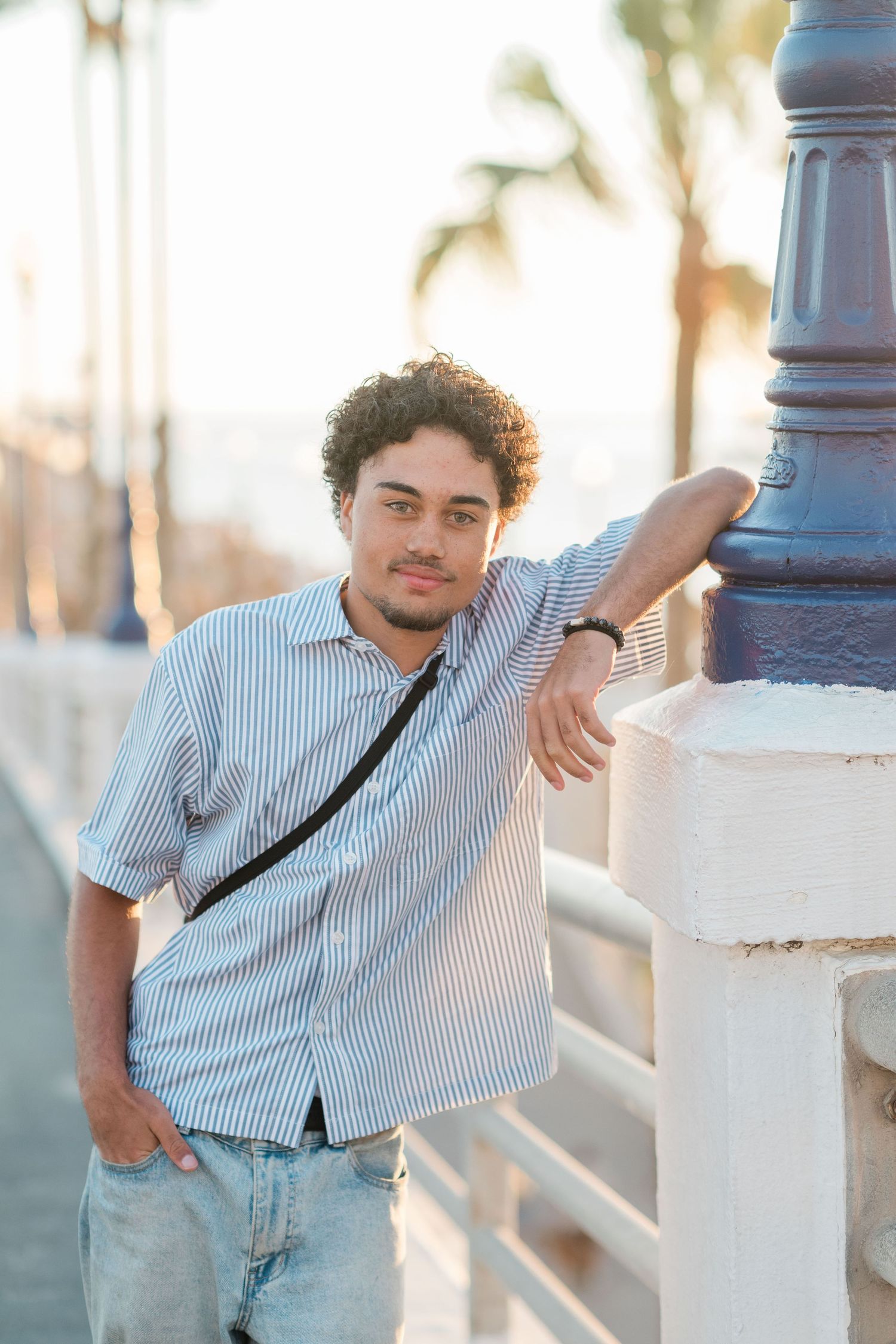 Young man in striped shirt leaning on white railing near a lamppost with palm trees in the background.