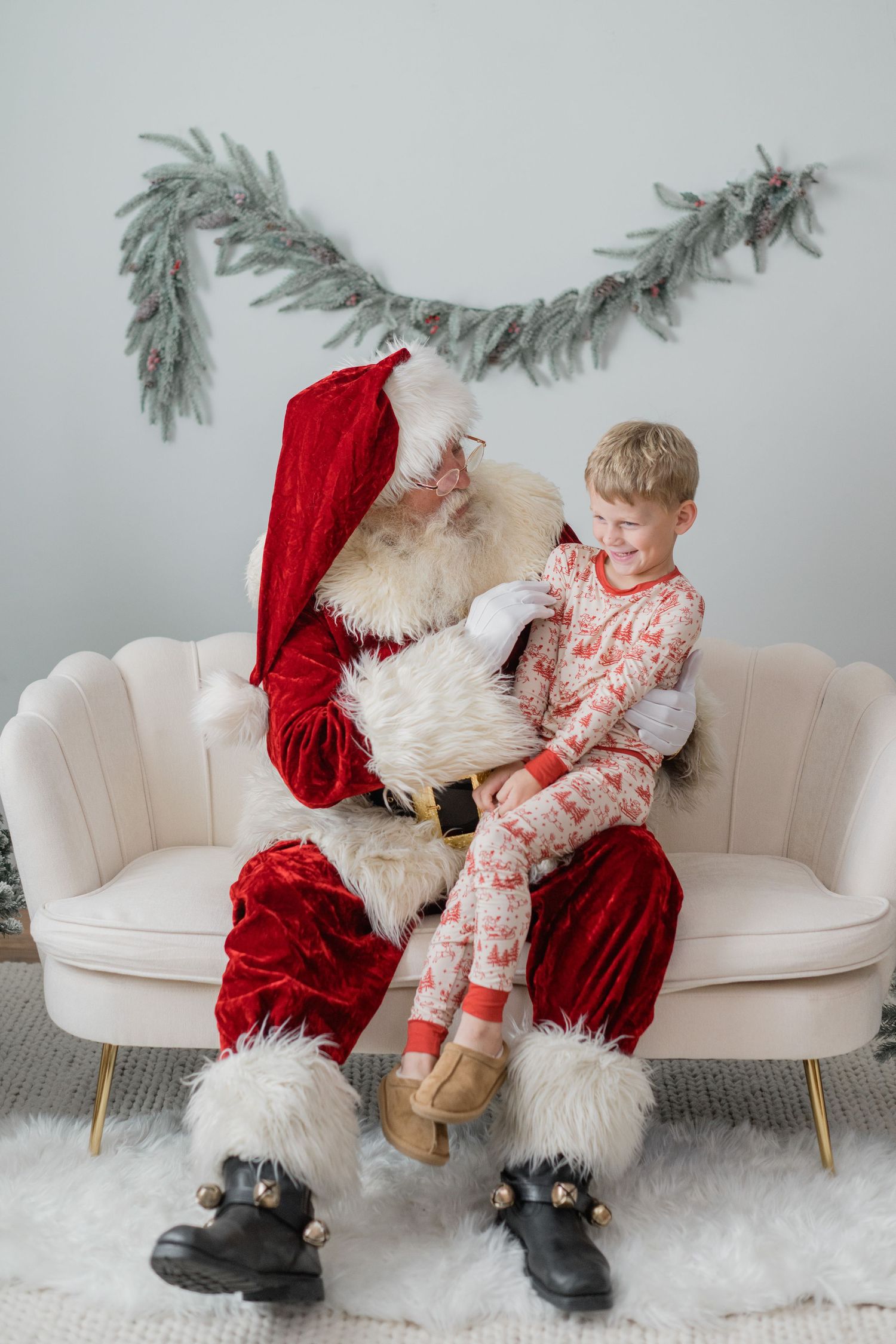 Young blonde boy in Christmas pajamas sits on Santa Clauss lap on a cream sofa with holiday garland backdrop.