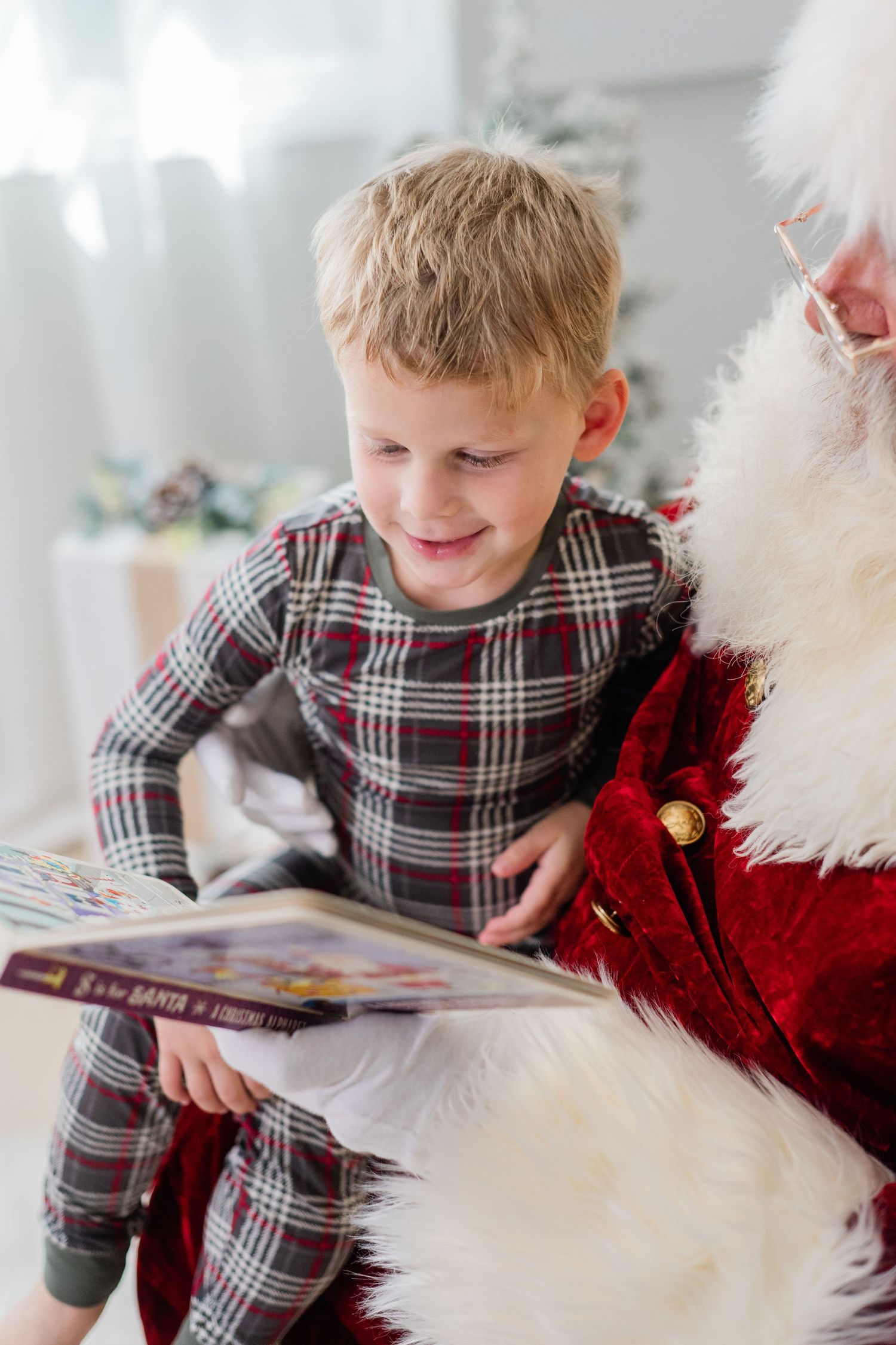 A young boy in plaid pajamas smiles while reading a Christmas book with Santa Claus during a holiday photo session.