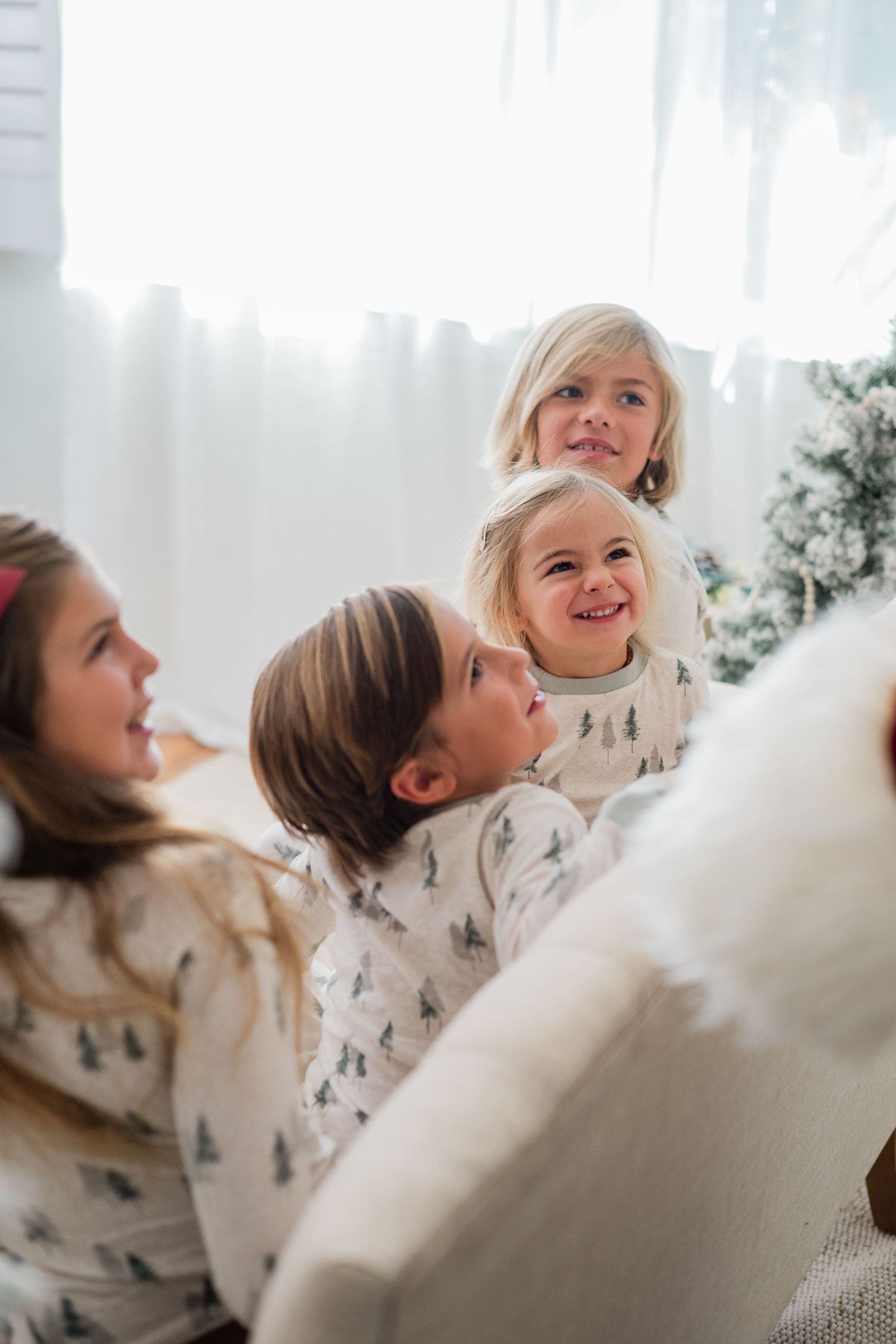 Four children in matching Christmas tree pajamas laughing and playing together near a decorated holiday tree.