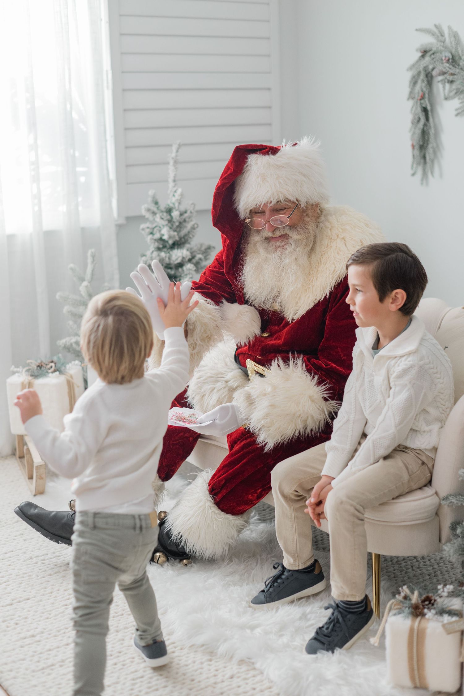 Santa Claus giving a high five to a young boy while another child sits beside him in a festive white Christmas room.