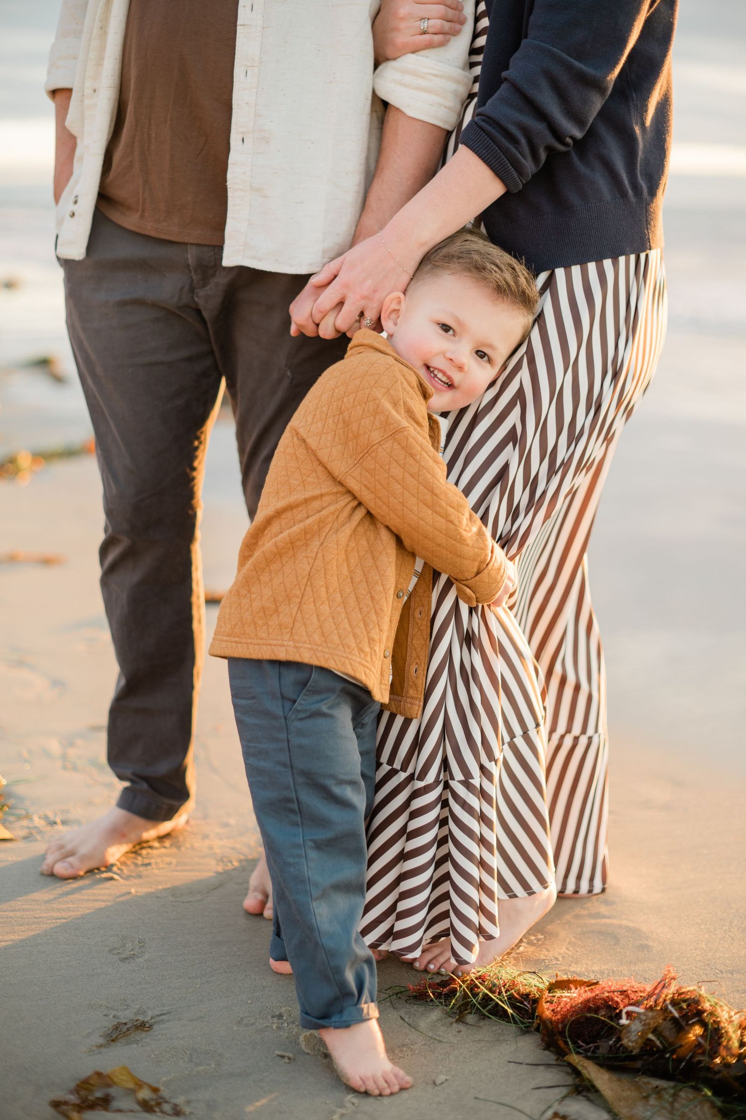 A smiling toddler boy in a mustard sweater hugs his mothers striped skirt on a sandy beach.