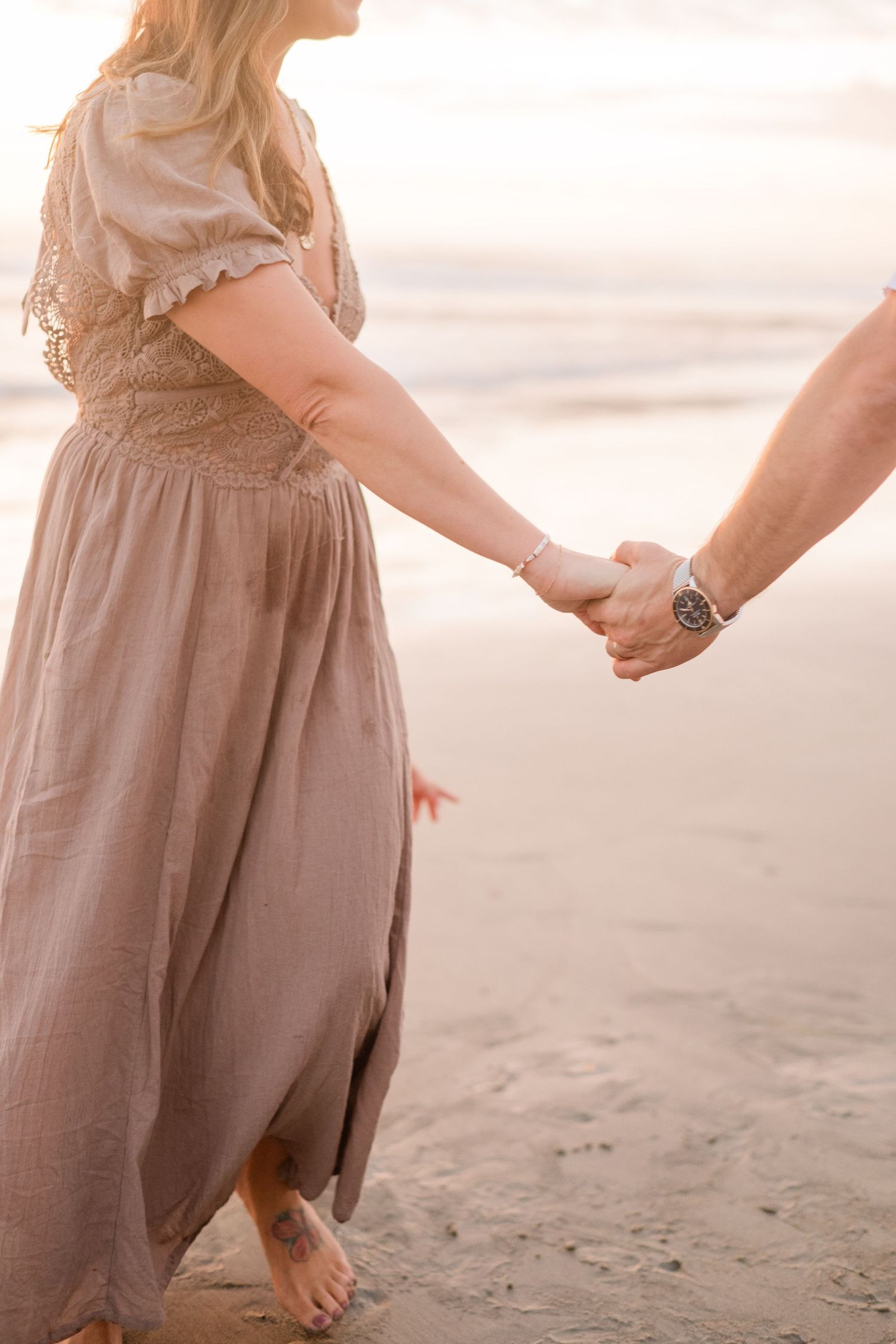 Couple holding hands on a sandy beach at sunset, woman in flowing maxi dress, man wearing a watch.