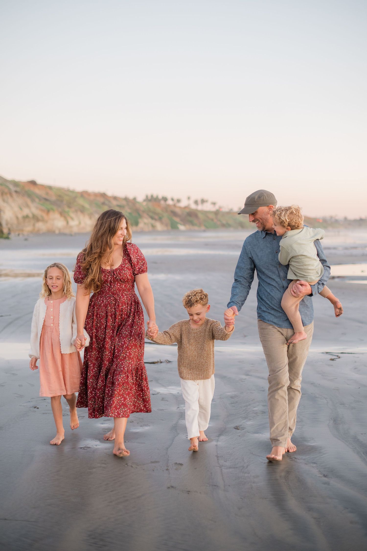 Family of five walking hand-in-hand on a sandy beach at sunset near rocky cliffs.