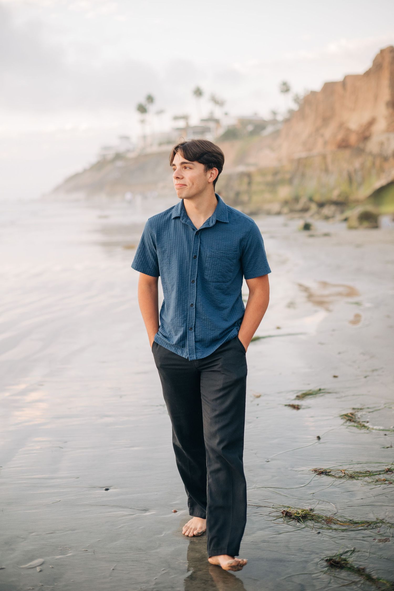 Young man in blue shirt and black pants walking barefoot on a sandy beach with coastal cliffs in the background.