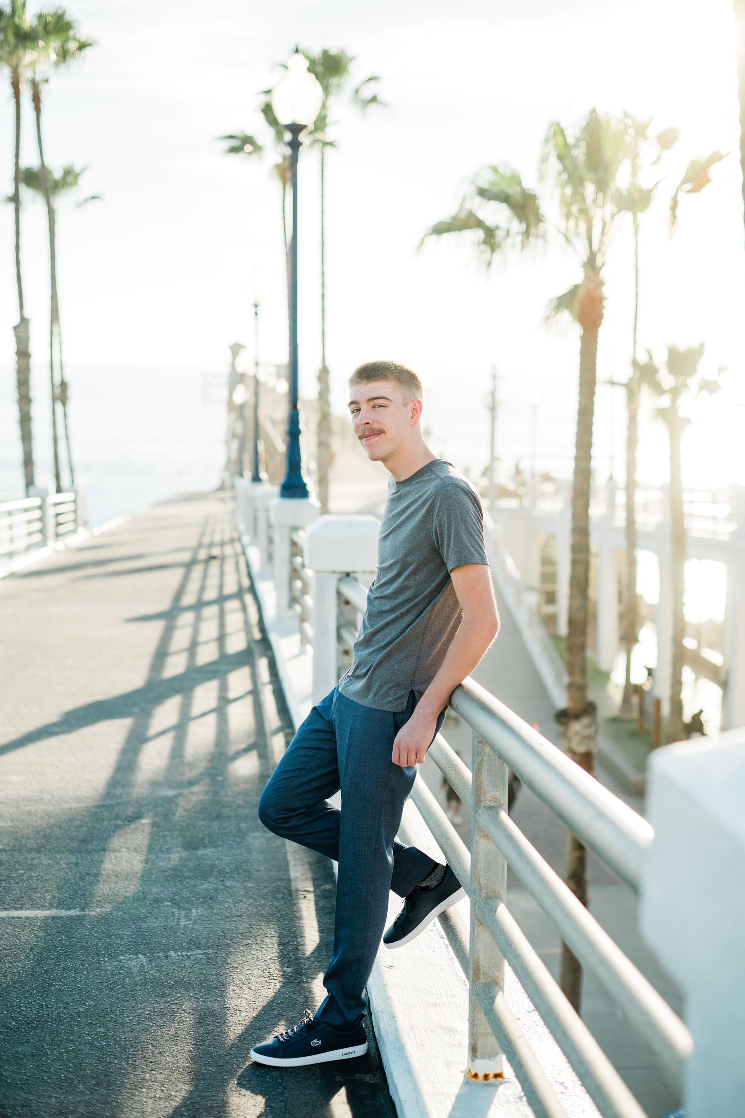 Young man in gray t-shirt and jeans leaning on white railing of sunny pier with palm trees in background.
